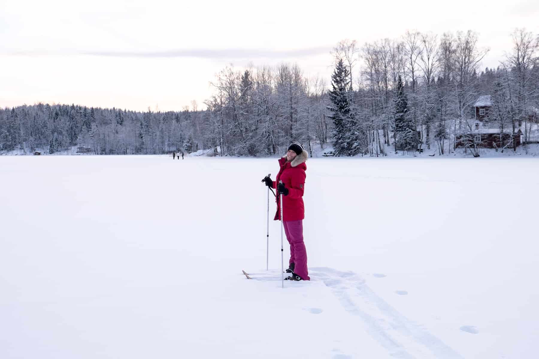 A woman in a red coat and pink ski pants cross country skiing on a frozen lake in Finland. Houses can be seen in the forest surrounding the lake. 