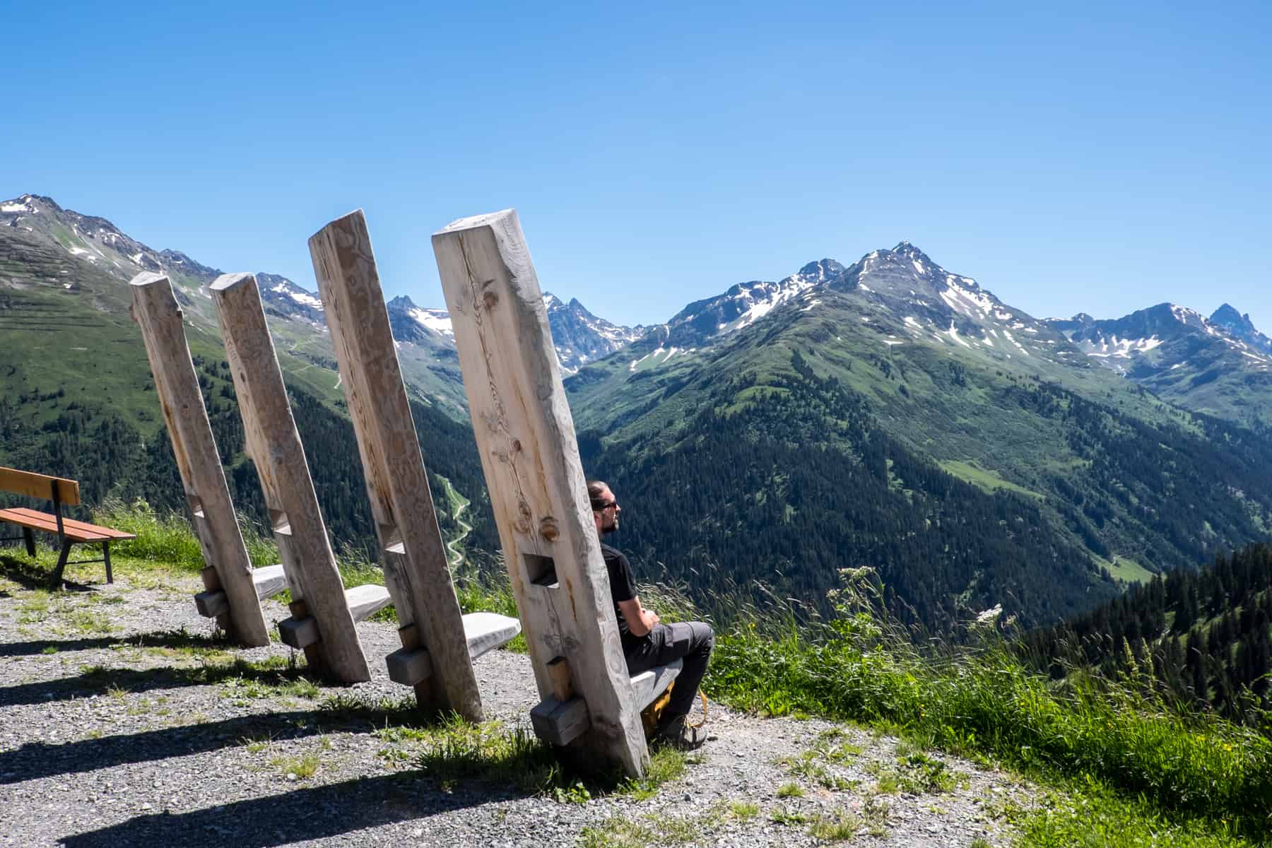A man sits on one of four tall chiars carved from trees trucks overlooking St. Anton am Arlberg in summer sun