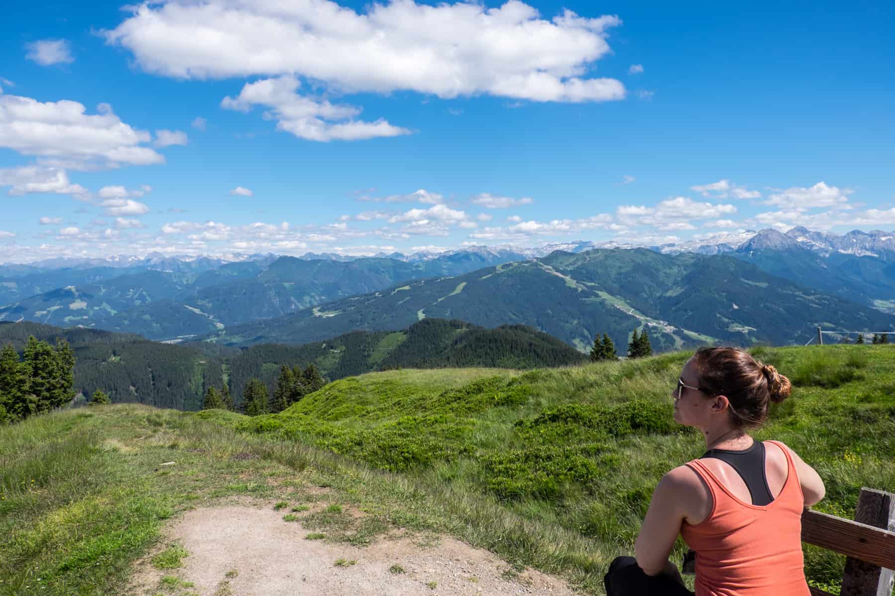 A woman in an orange vest sits on a bench admiring the panoramic Austrian Alps view from atop the Hochgründeck Mountain