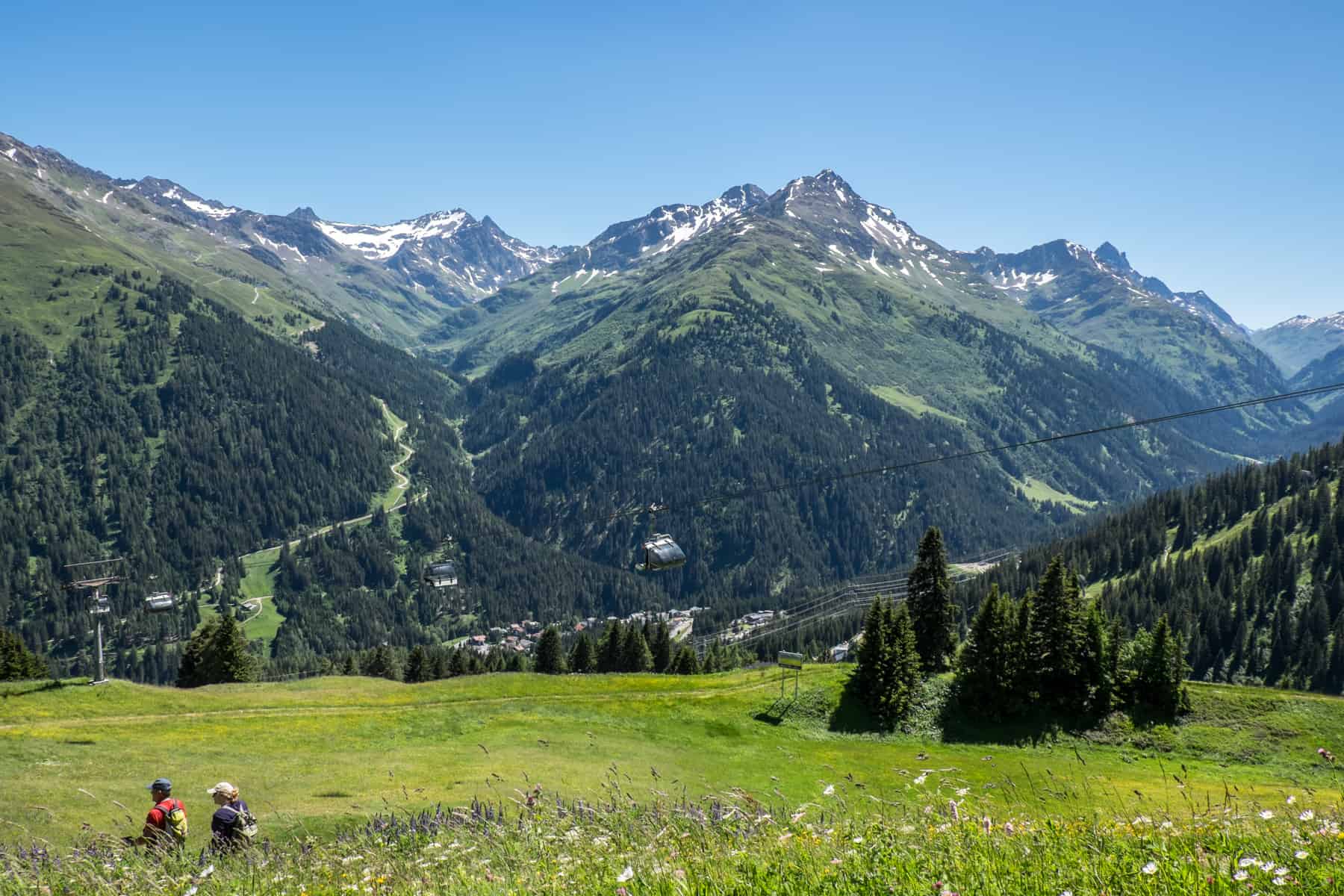 Two hikers start their descent on a green mountain trail in St. Anton am Arlberg with the jagged Austrian Alps the background