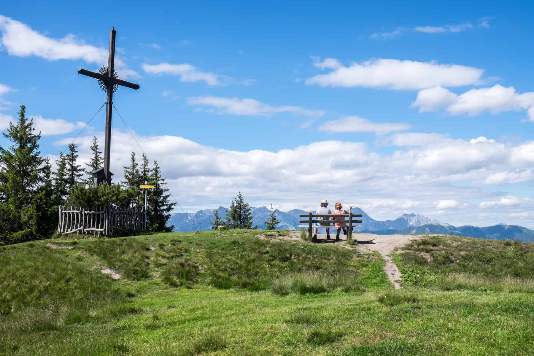 A couple sit on a bench next to the hiking cross on the green summit of Hochgründeck Mountain in Austria