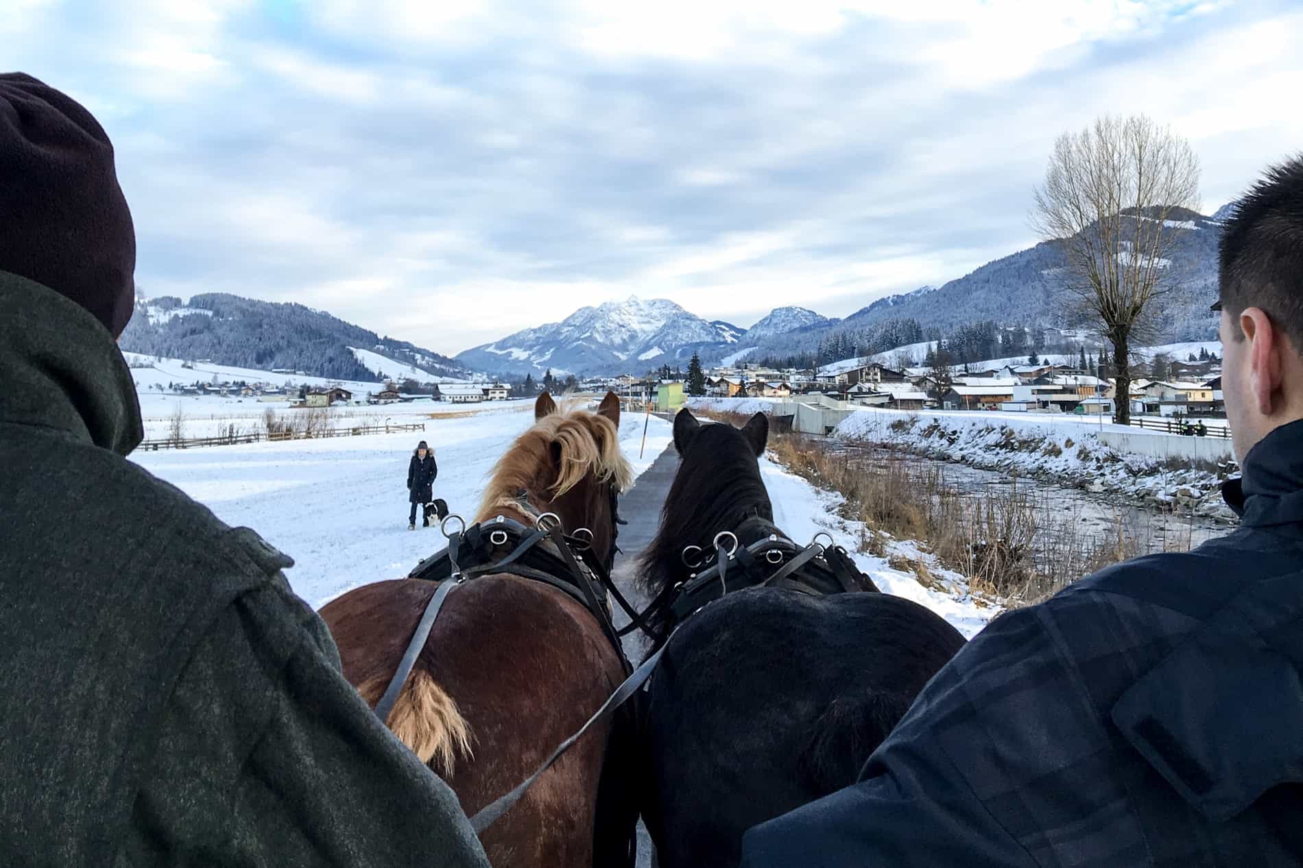 Two people ride a horse and cart through a snow coated Austrian village in winter. 