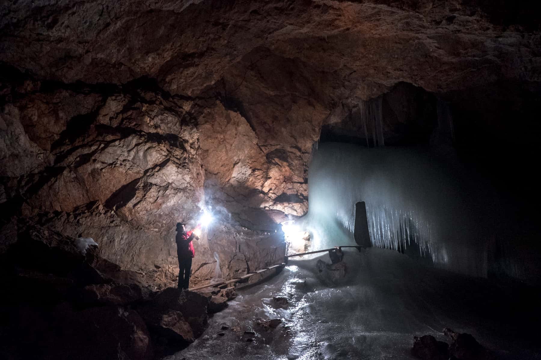 A guide in a red coat, holding a sparking light stands with a cave system next to a sheet of blue ice in Eisriesenwelt, Austria