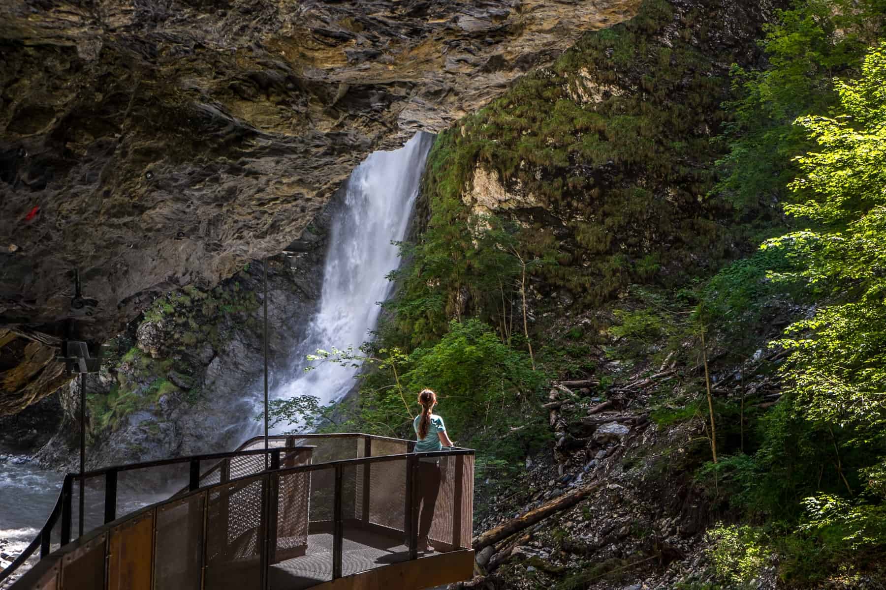 A woman in a blue t-shirt stands on an iron viewing platform looking towards a gushing waterfall in the Liechtensteinklamm gorge in St. Johann im Pongau Austria