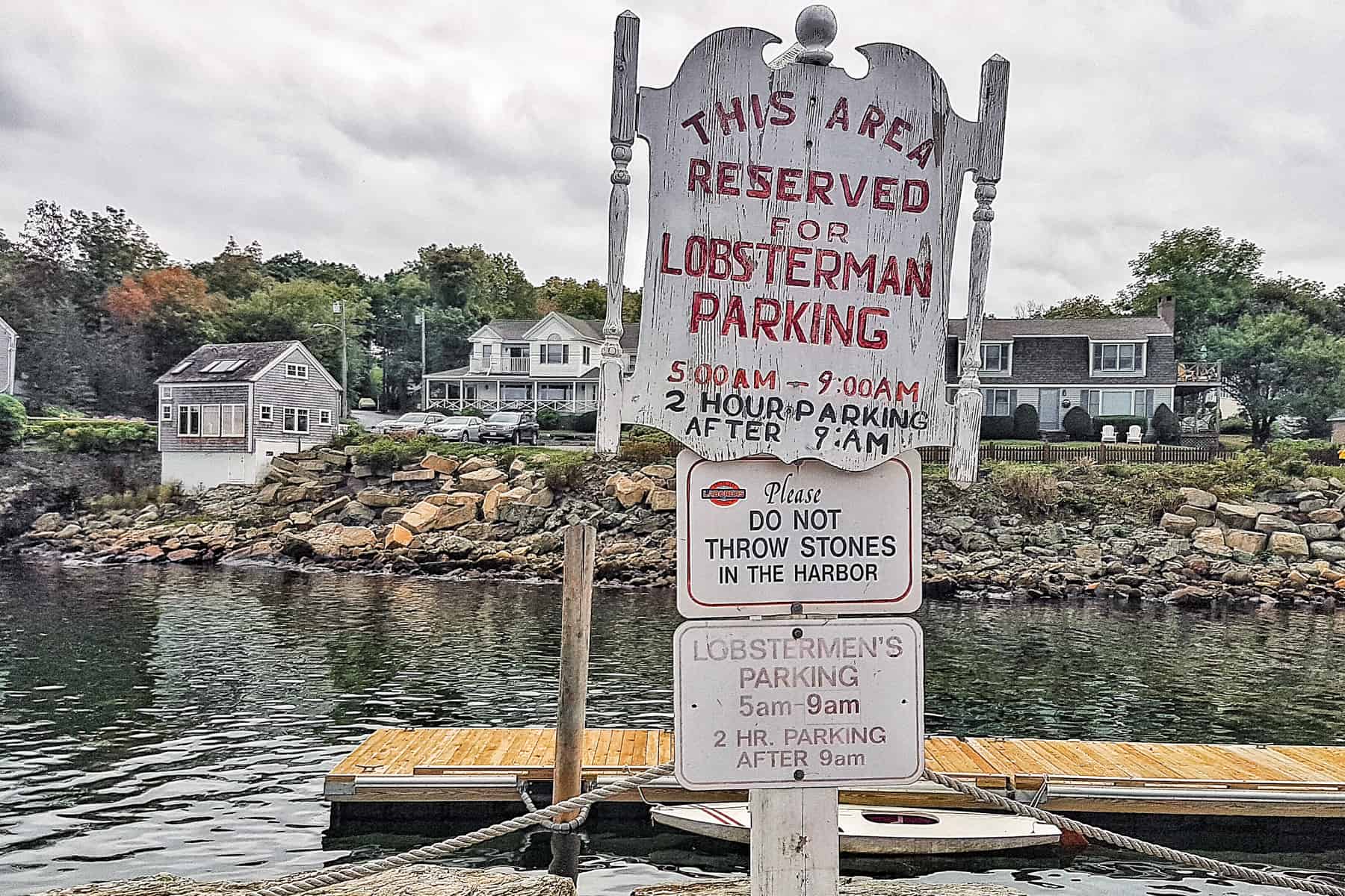 A white wooden sign in Maine that reads: This area is reserved for lobsterman Parking. Behinds are white houses next to the rocky shoreline. 