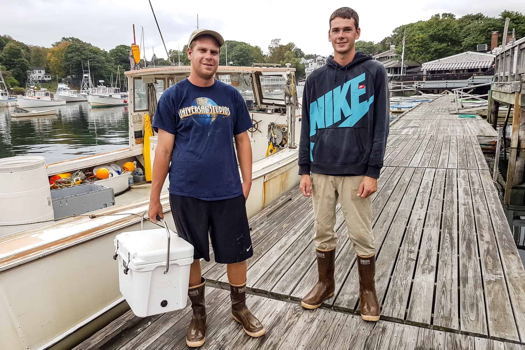 Two young Lobster fishermen in Maine next to a boat. One carries a white cooler box, full of fresh fish. 