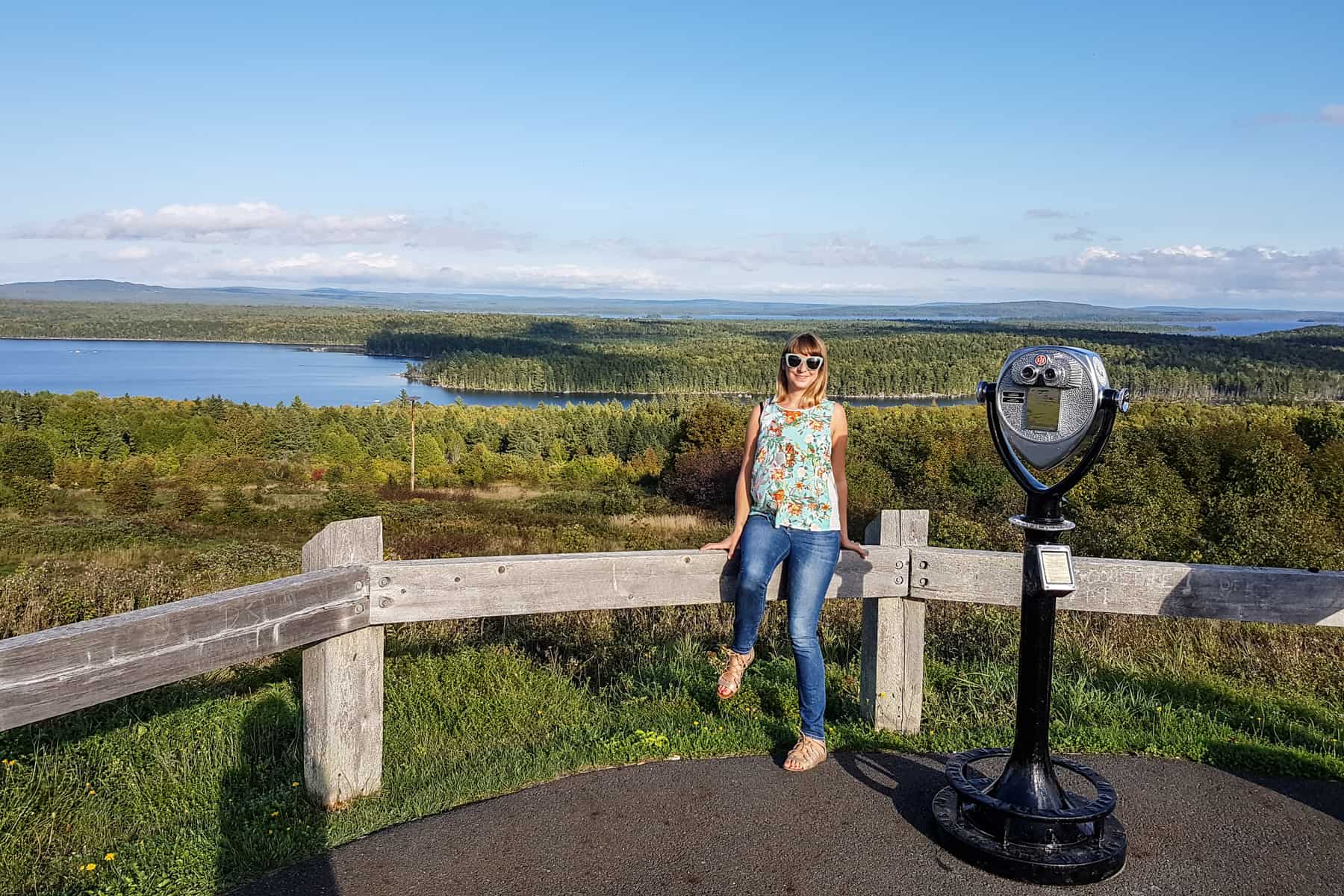 A blonde woman sits on a wooden fence next to a telescope viewer, with a background of green islands and waterways.