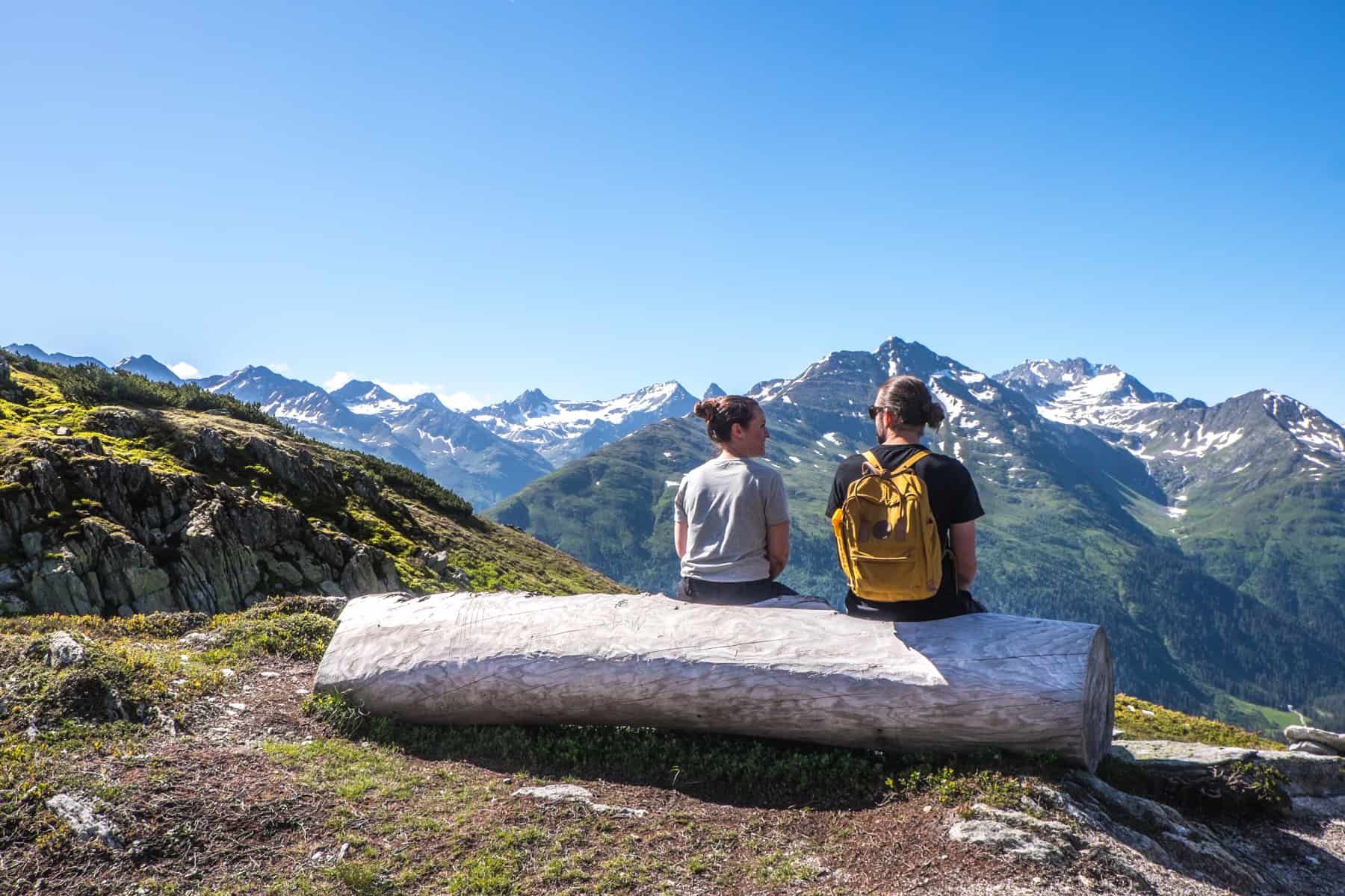 A woman and a man sit on a white tree truck shaped into a chair, overlooking the Austrian Alps from a mountain in St Anton