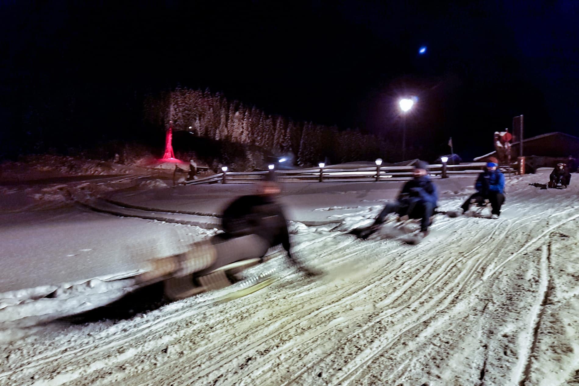 Four people glide fast on the snow on a toboggan at nighttime. 