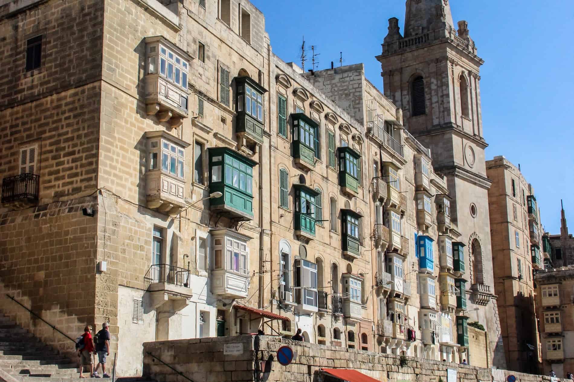 A long yellow-stone building in Valletta, whose facade is dotted with cream, green and blue-painted balconies. 