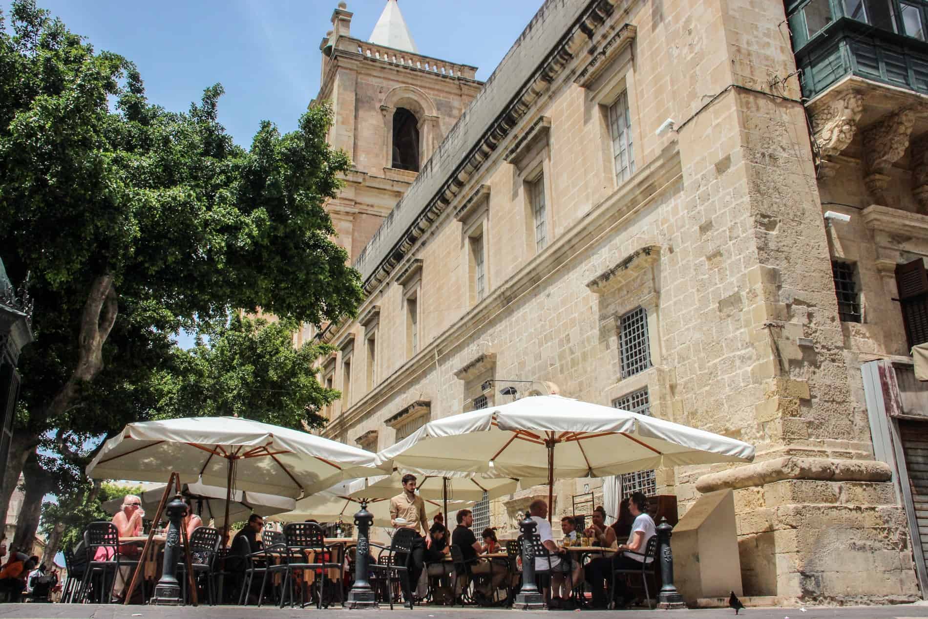 People dining at an outdoor cafe in Valletta in front of a grand limestone building with a tower. 