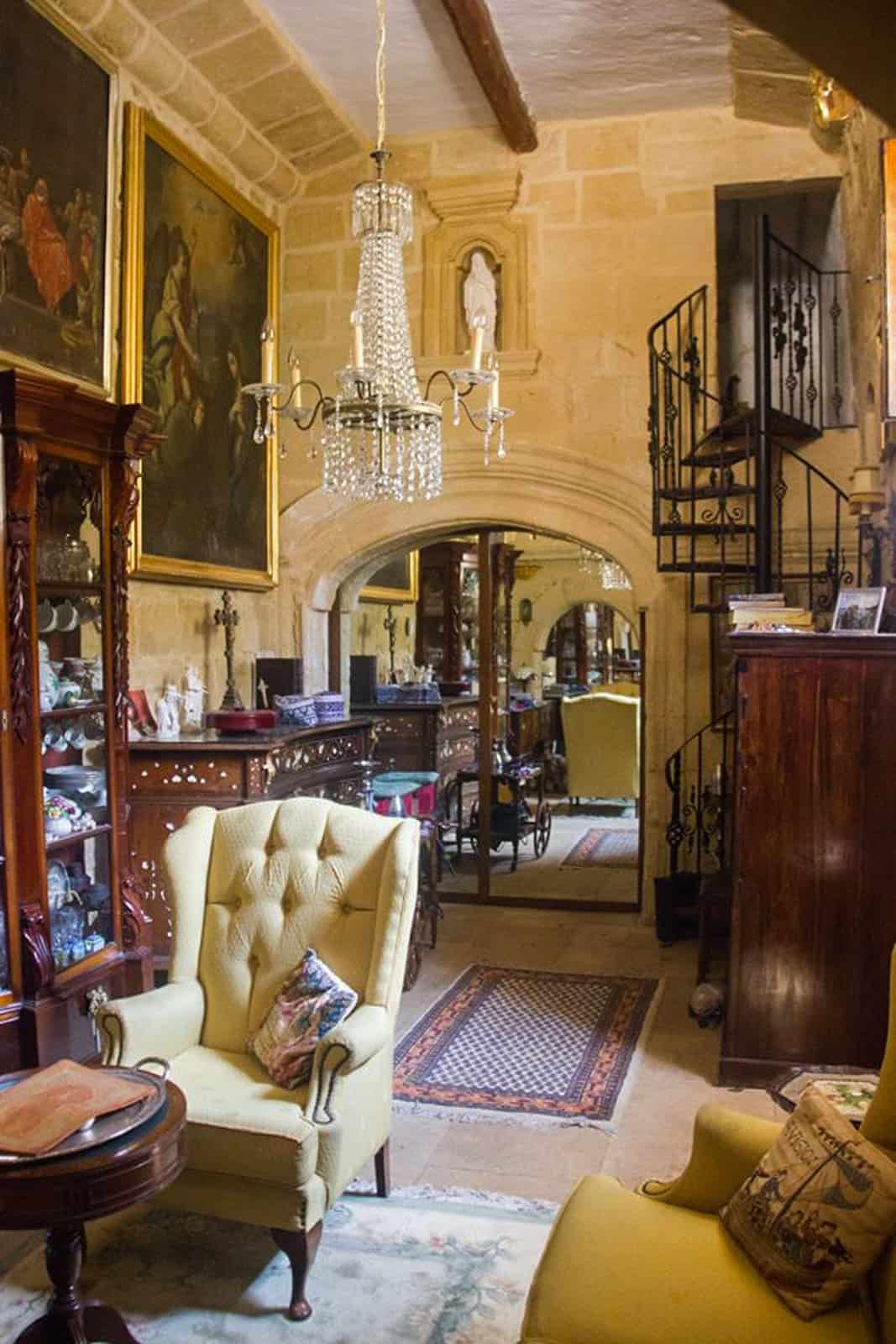 A limestone wall chapel room with a black spiral staircase filled with dark wood furniture and plump armchairs. 