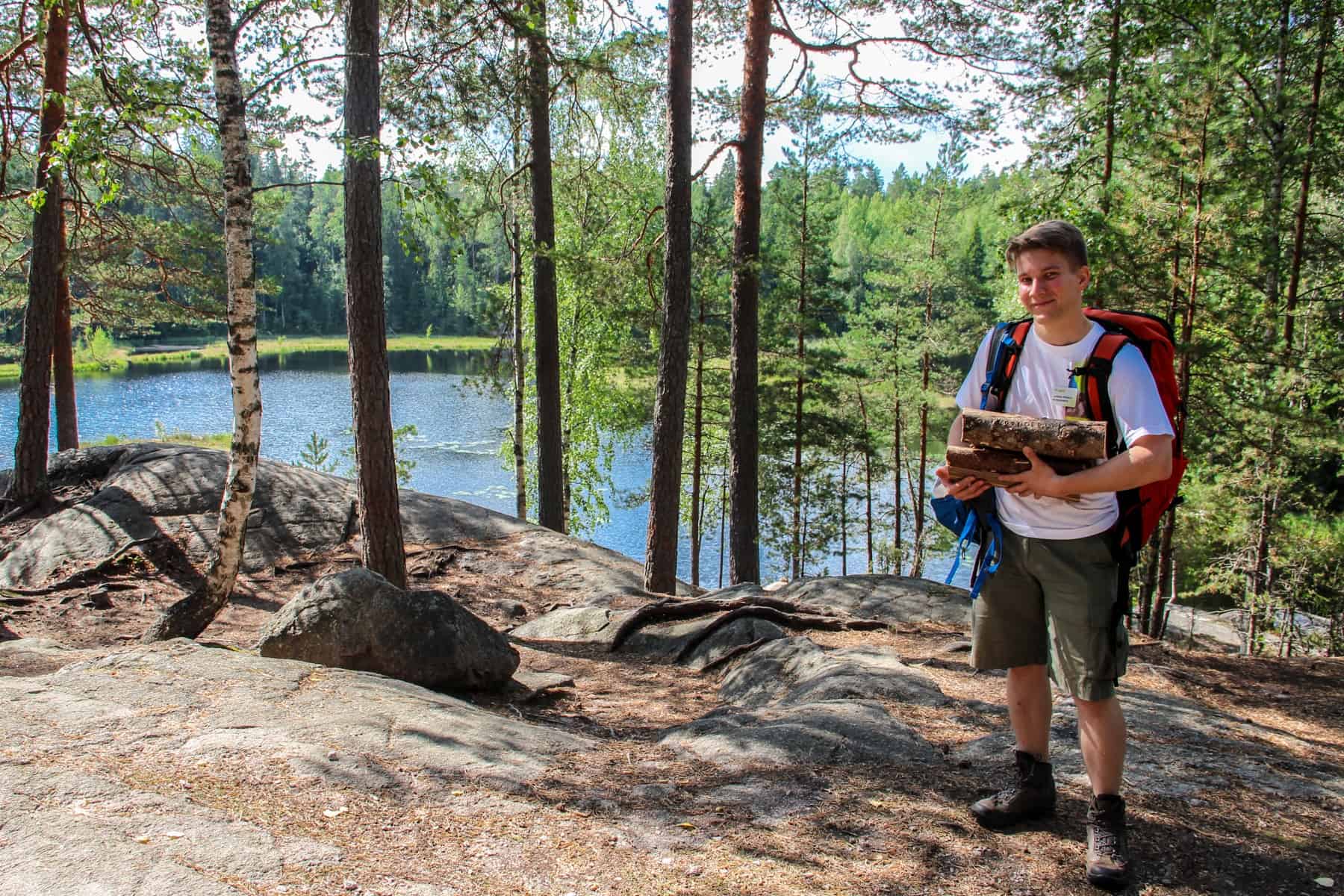 A man with a red backpack carries two, small tree logs on a forest path next to a lake in Finland. 