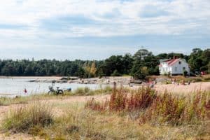 A dry grass covered beach next to the sea in Finland, backed by a white house with an orange roof in front of dense forest area.