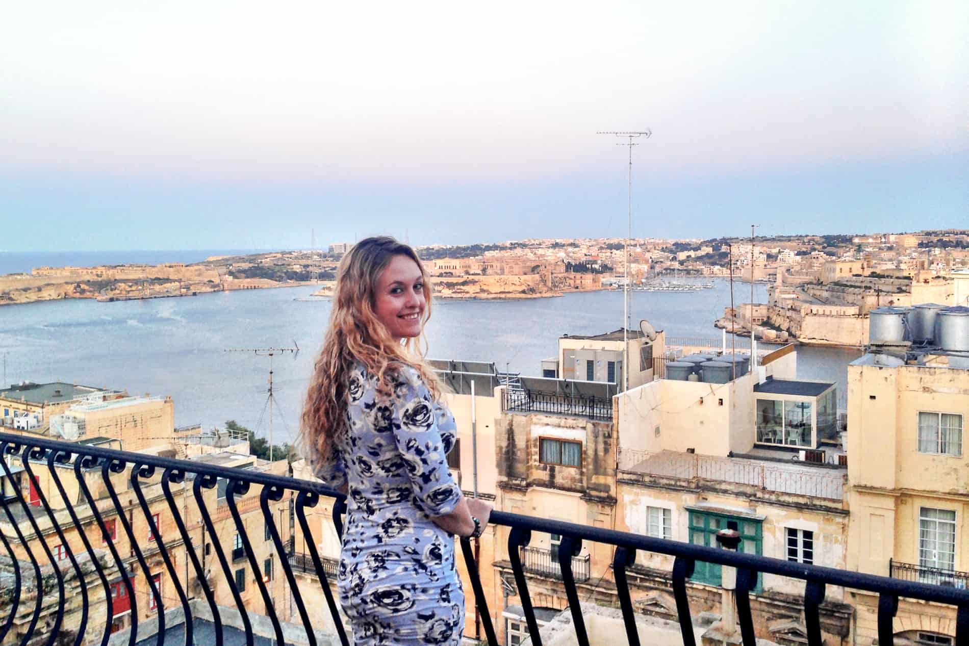 A woman smiles at the camera, standing at a Maltese Palazzo balcony overlooking a bay lined with yellow limestone buildings. 