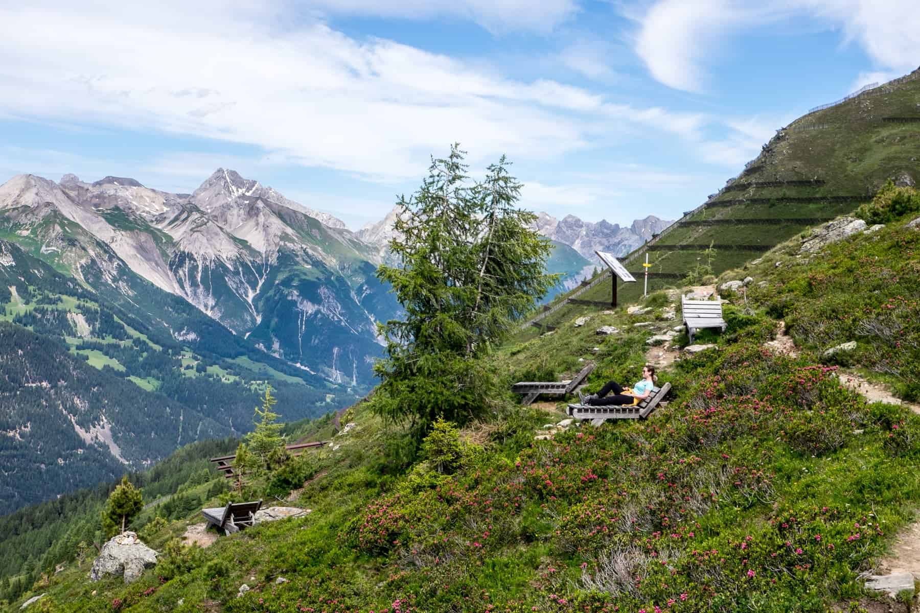 A woman rests on a wooden chair on a mountain slope, surrounded by flowers, on the St.Anton Alpenrosenweg hike