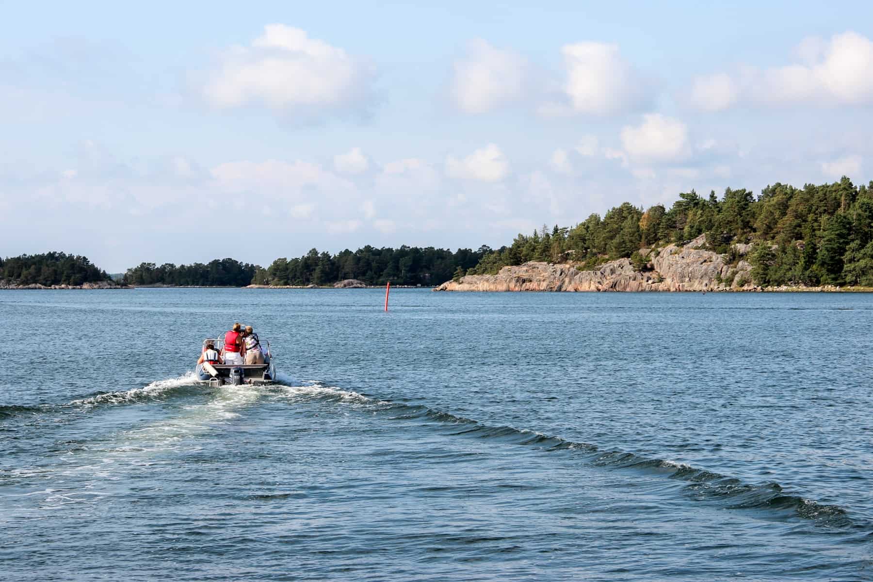 A small motorised boat with four people moves through a wide body of silver-blue water towards low rock formations covered in forest.