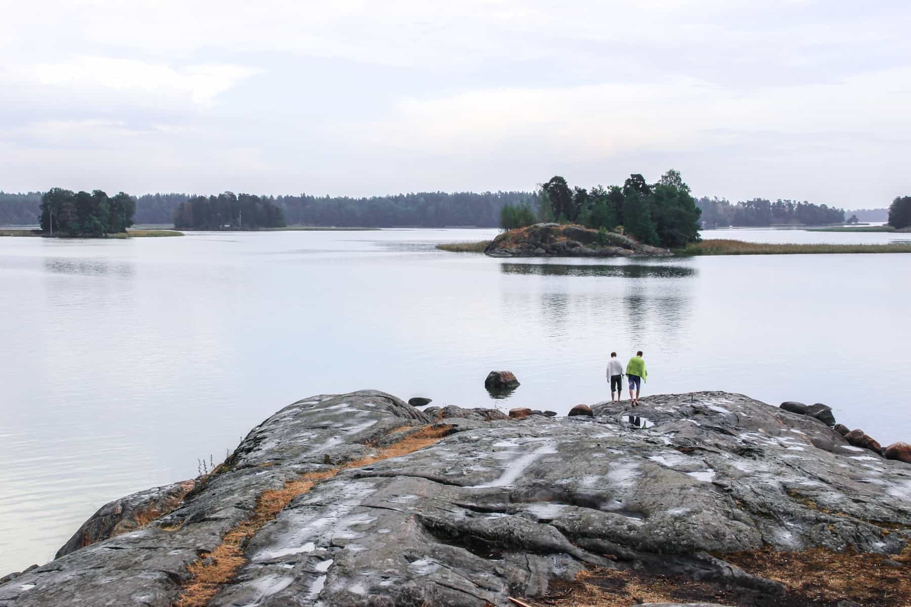 Two people stand upon a large silver rock formation covered in orange moss. They look out towards smaller, tree-covered islands, in the Southern Finland Archipelago. 