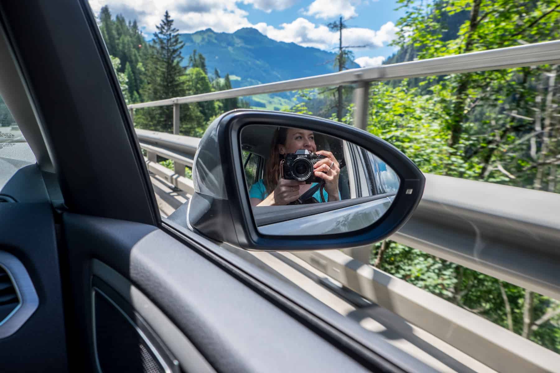 The reflection of a woman holding a camera seen in a car's side mirror. Green forest and mountains can be seen outside in the background