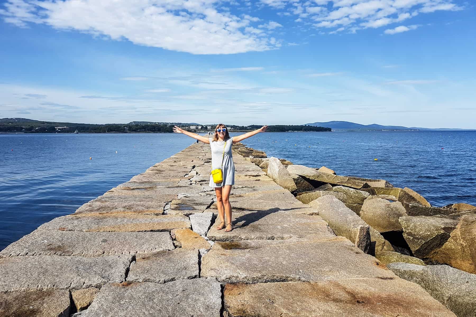A woman in a grey dress and yellow handbag stands on a long seaport pier made of large stones in Camden, Maine. 