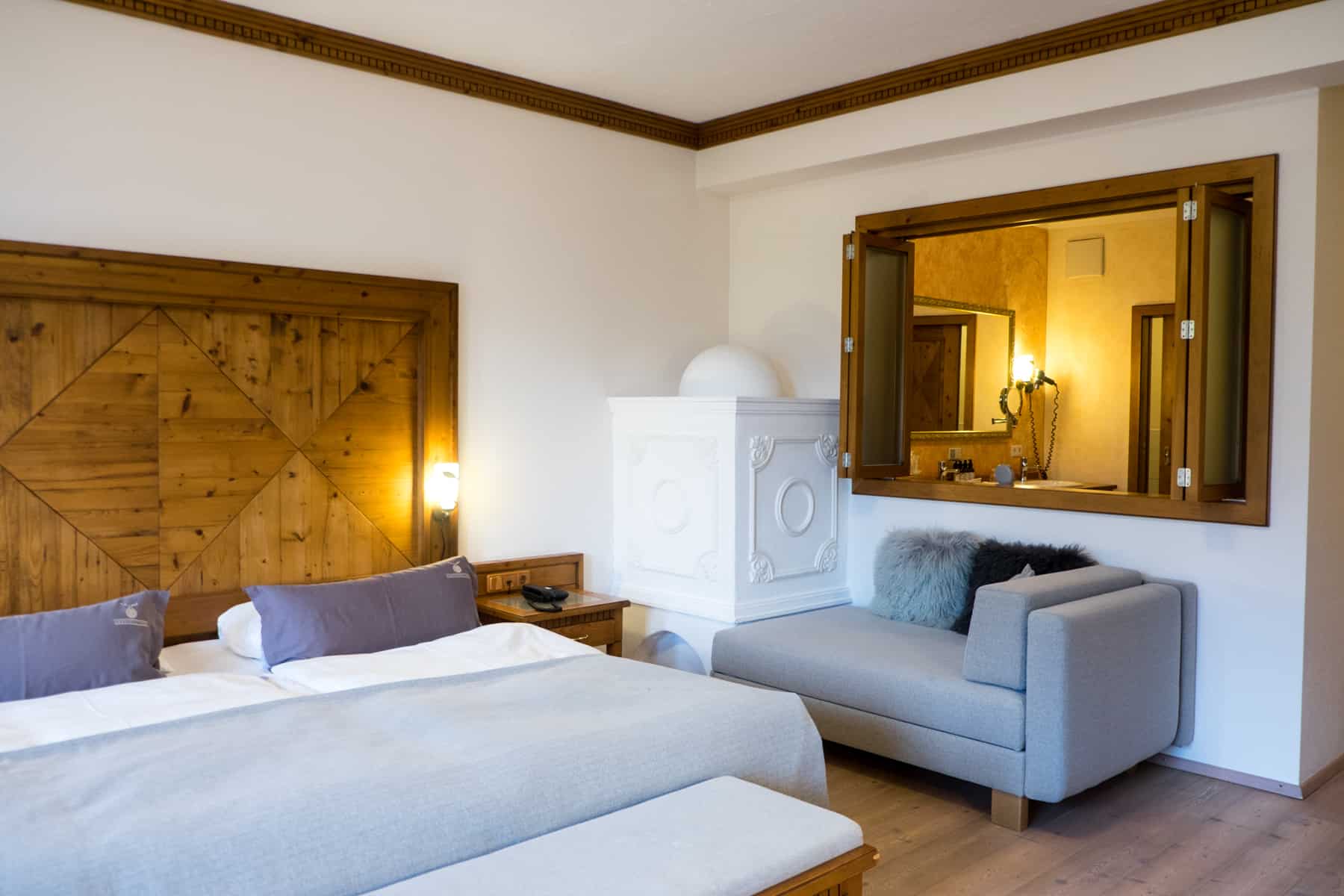 The classic, wooden trim and white-grey minimalist style interior of a bedroom at the Alpendorf Hotel Oberforsthof, with an open wall view to the bathroom