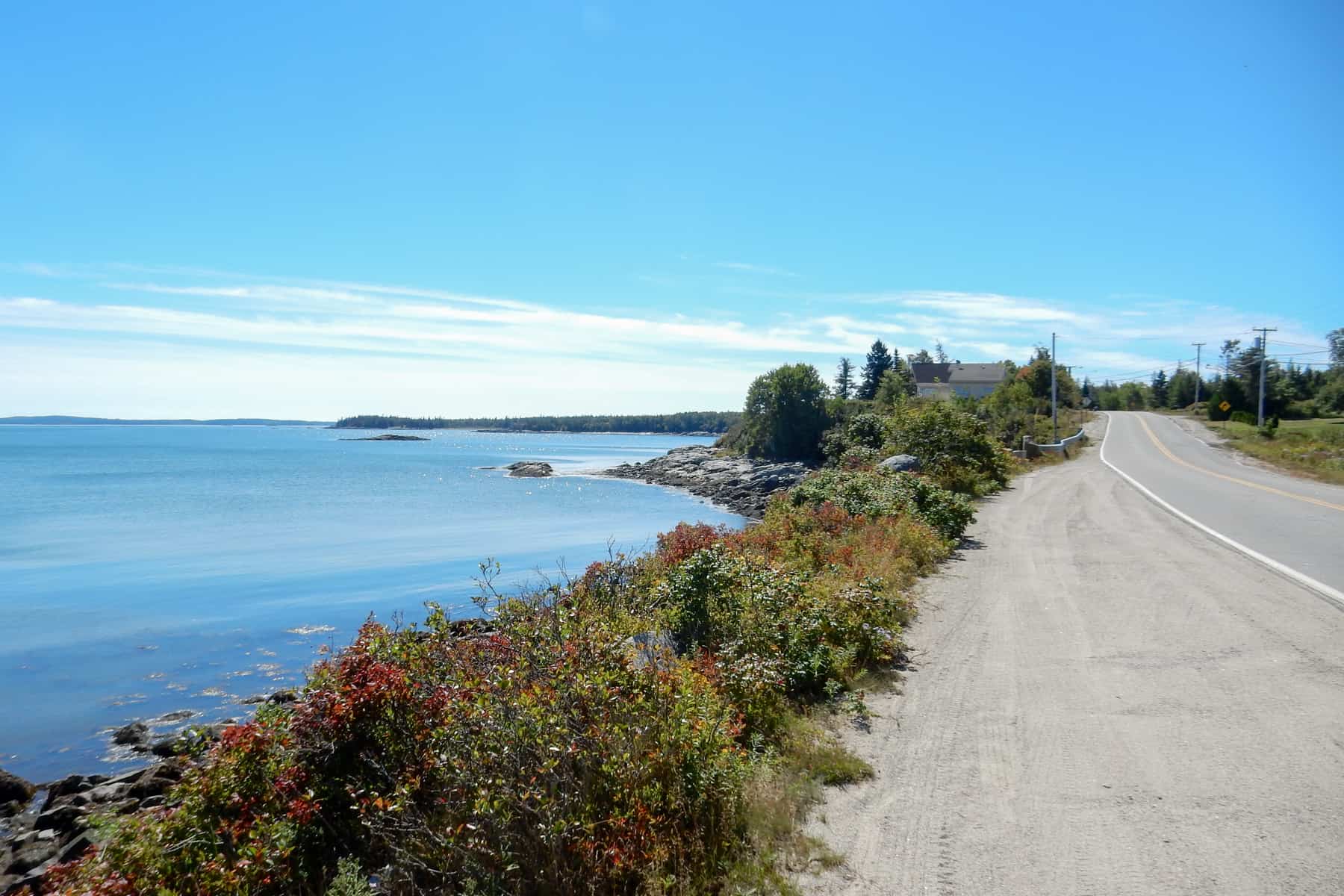 A long road lined with green and red foliage runs alongside the wide ocean - know as the Route 187 Bold Coast Scenic Byway in Maine.