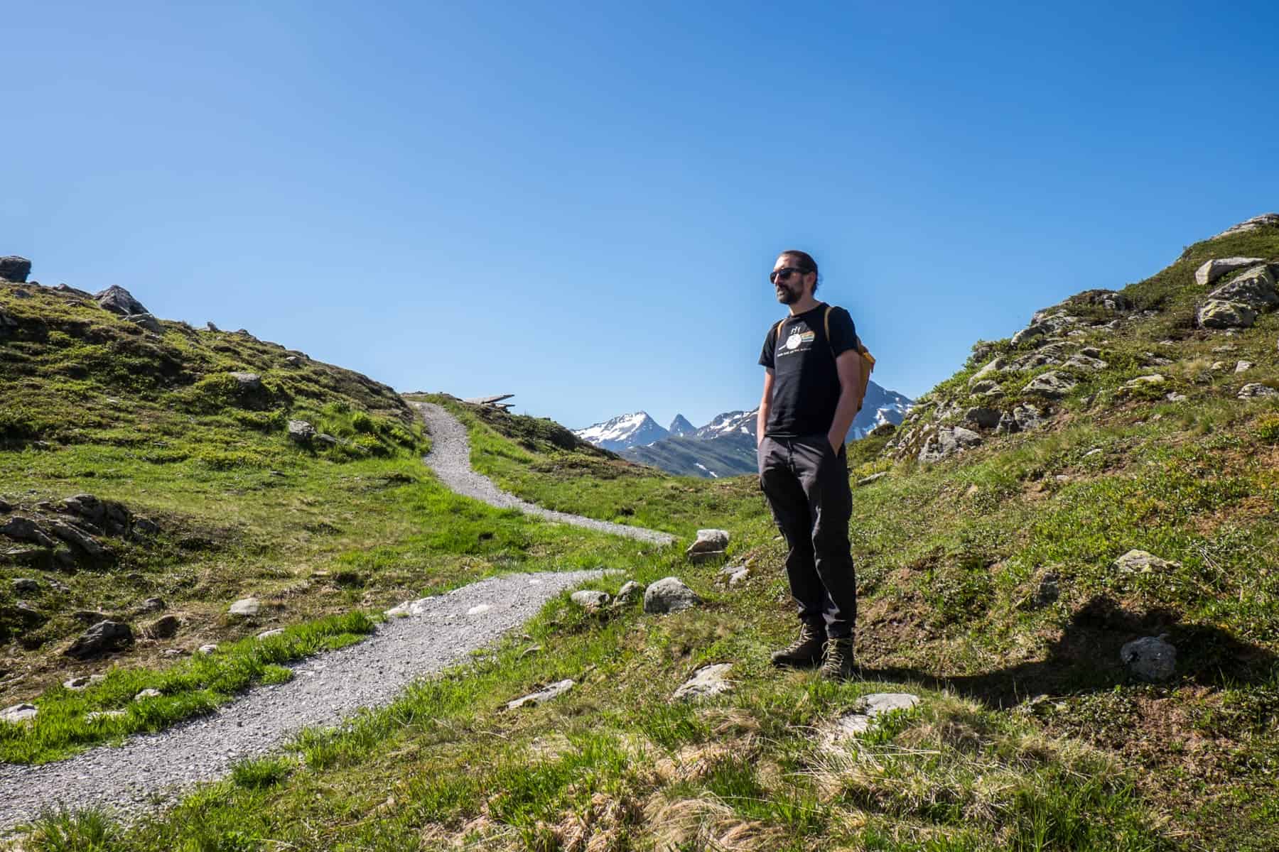 A man stands on the grass next to a grey, gravel hiking trail in the mountains in St Anton am Arlberg in Austria