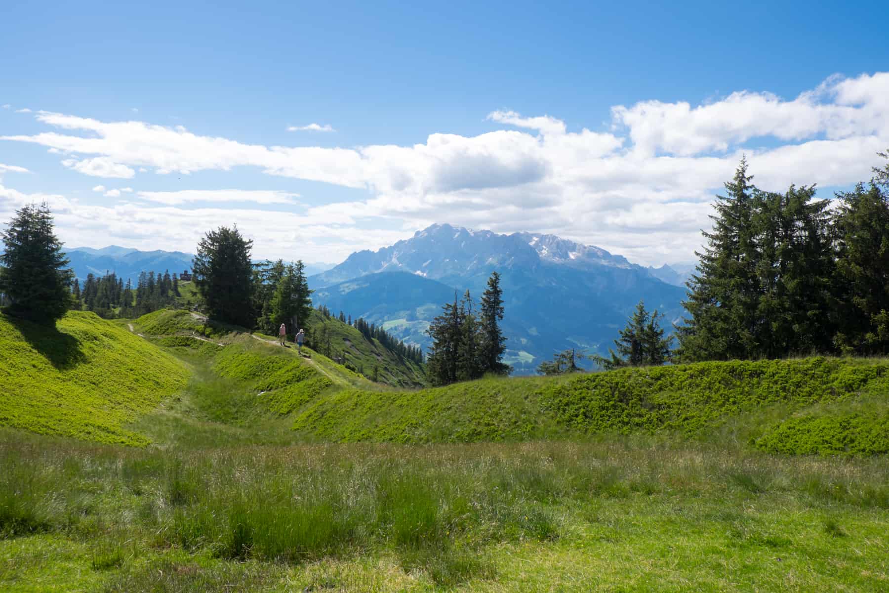 Two people walk along a narrow track on a green ridge at the top of the Hochgründeck Mountain in Austria in summer