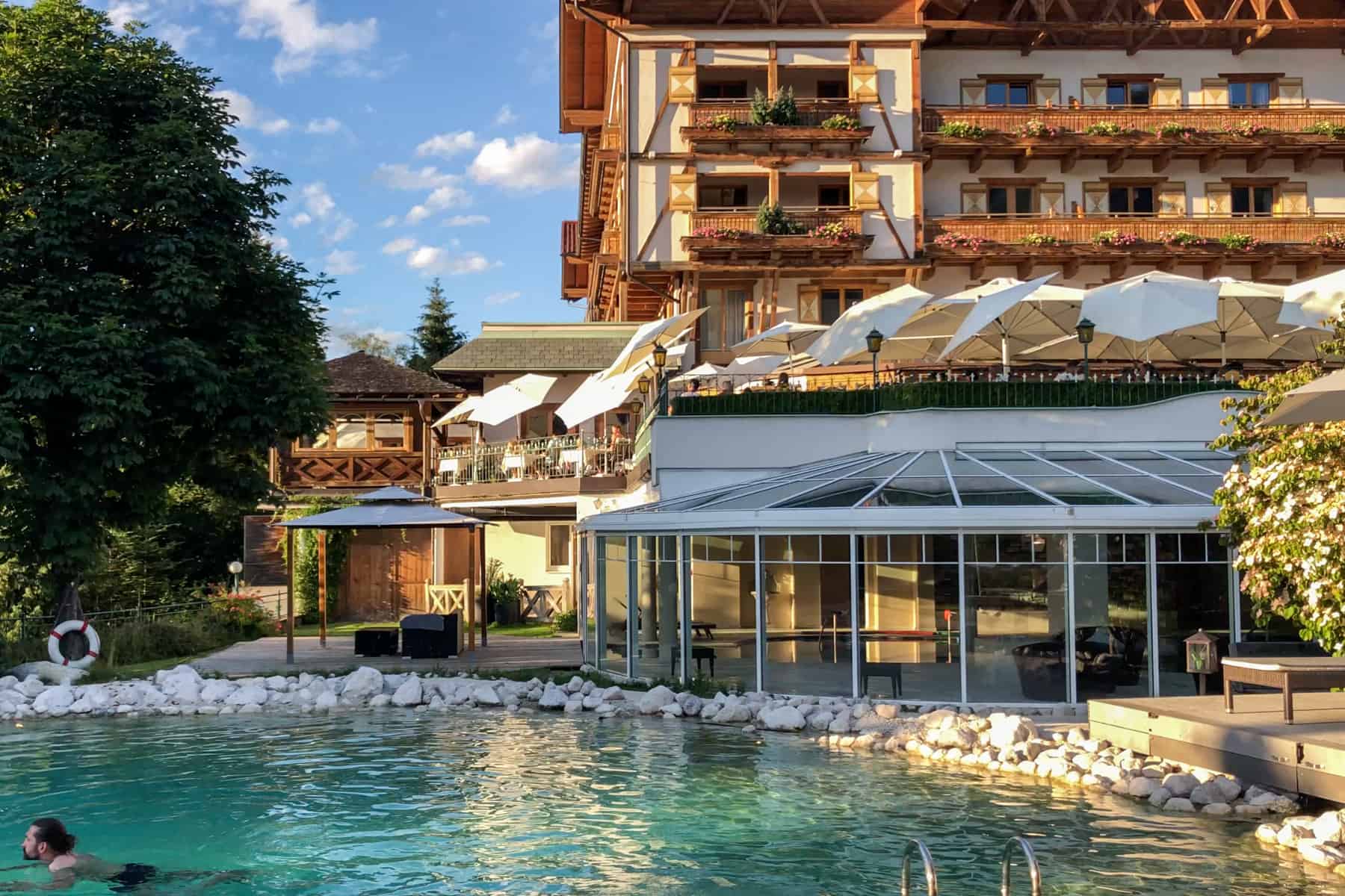 The wooden chalet-style exterior, umbrella level seating area in the middle, and a man swimming below in the blue-green swimming pool of the Alpendorf Hotel Oberforsthof in St. Johann im Pongau, Salzburg, Austria