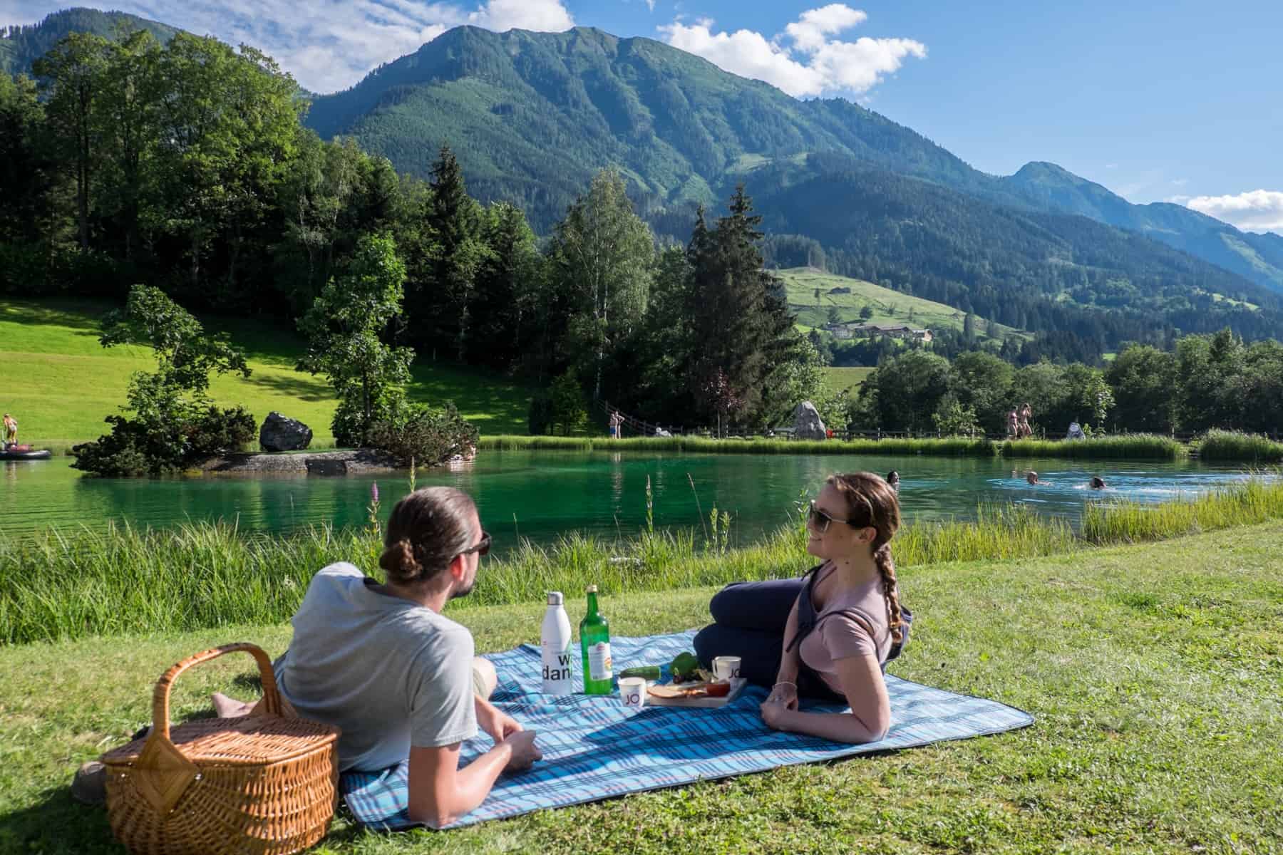 A man and woman look at one another while relaxing on a blue rug with a picnic next to the bathing lake, Badesee Plankenau in St. Johann im Pongau, Austria