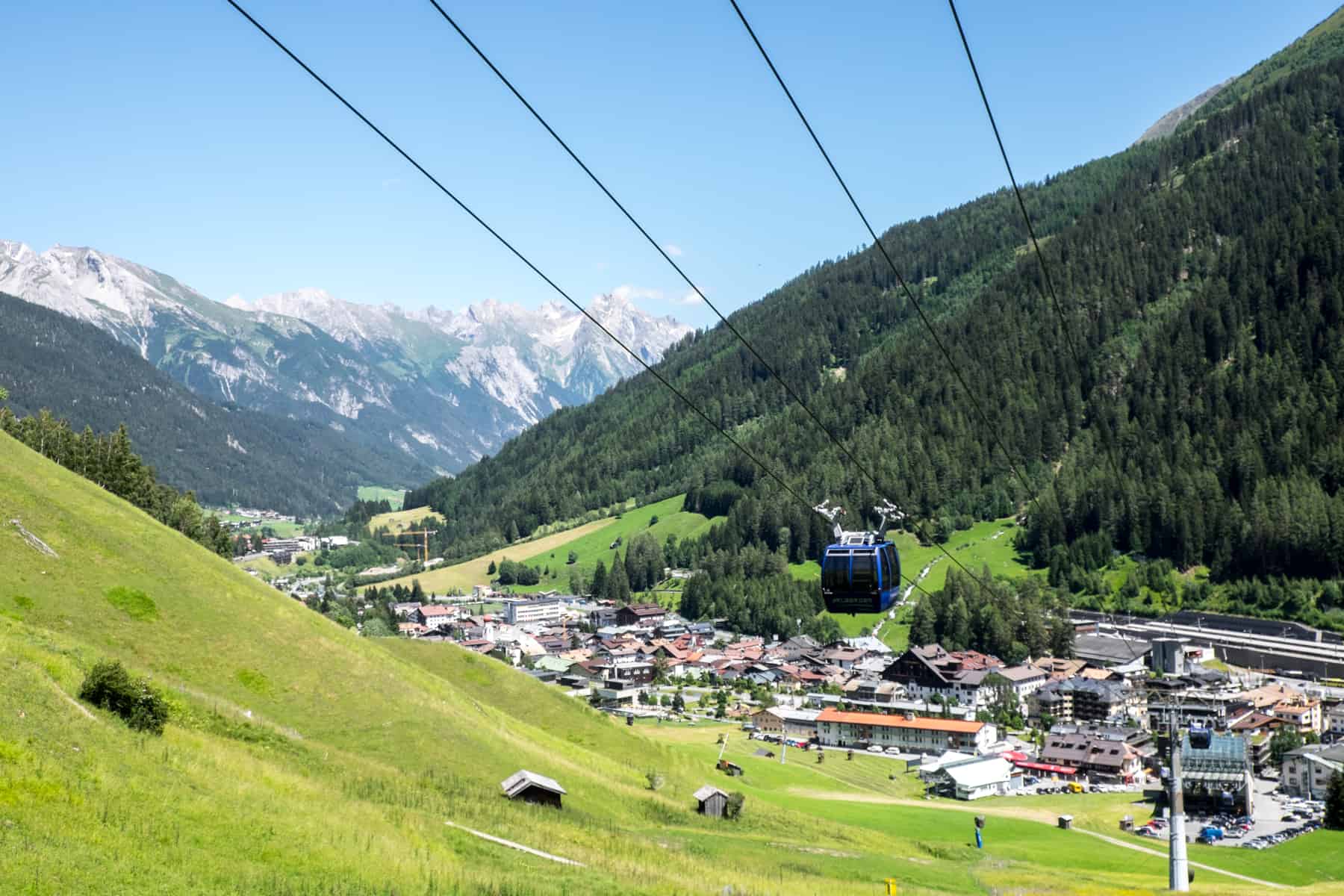 A blue cable car glides over a green valley from the cludter of buildings below - the village of St Anton in summer