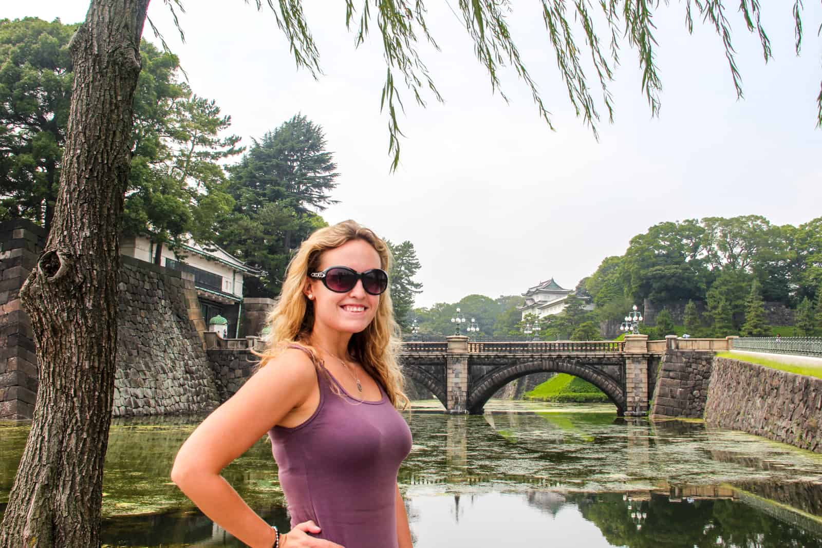 A woman stands in front of a lake with a stone bridge inside the Imperial Palace in Tokyo. 