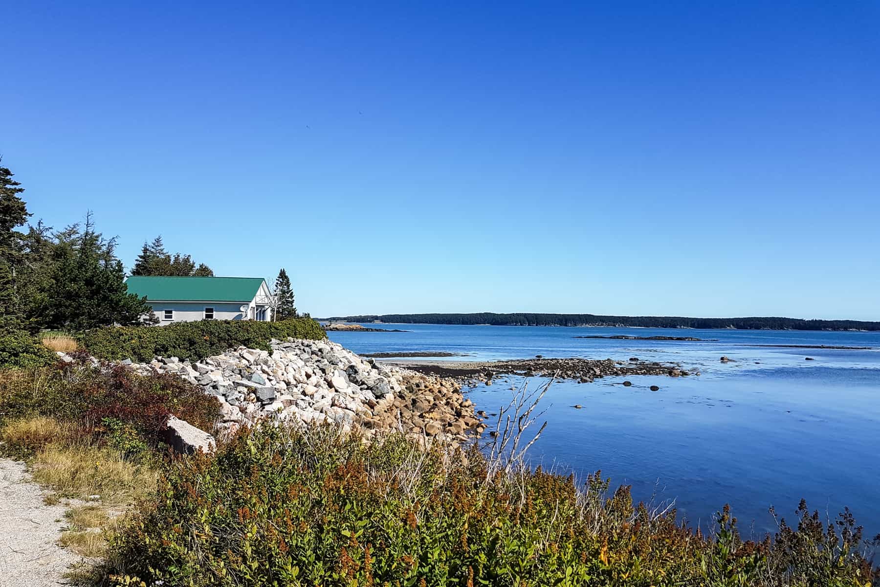 A white house with a mint green roof, stands solitary on a rugged ocean coastline - part of the Bold Coast Scenic Byway on the US Route 1 highway.