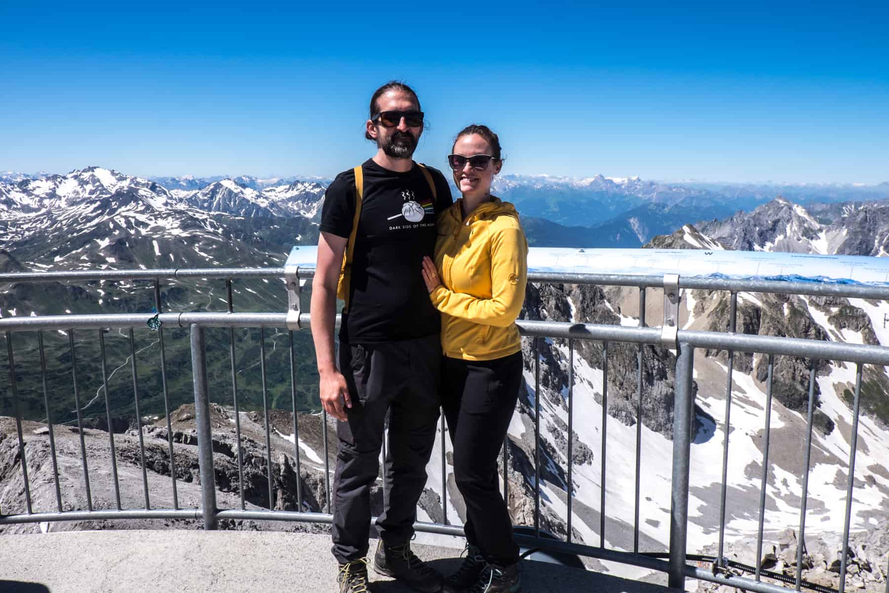 A man and woman hug standing on a mountain viewing platform in St Anton in Austria. In the background is a panorama of snow-capped Alps mountains
