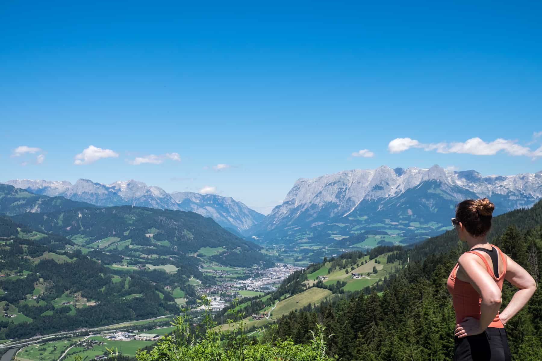 A woman in an orange vest looks out over a green valley filled with St. Johann im Pongau city, thats backed by flat-top mountains