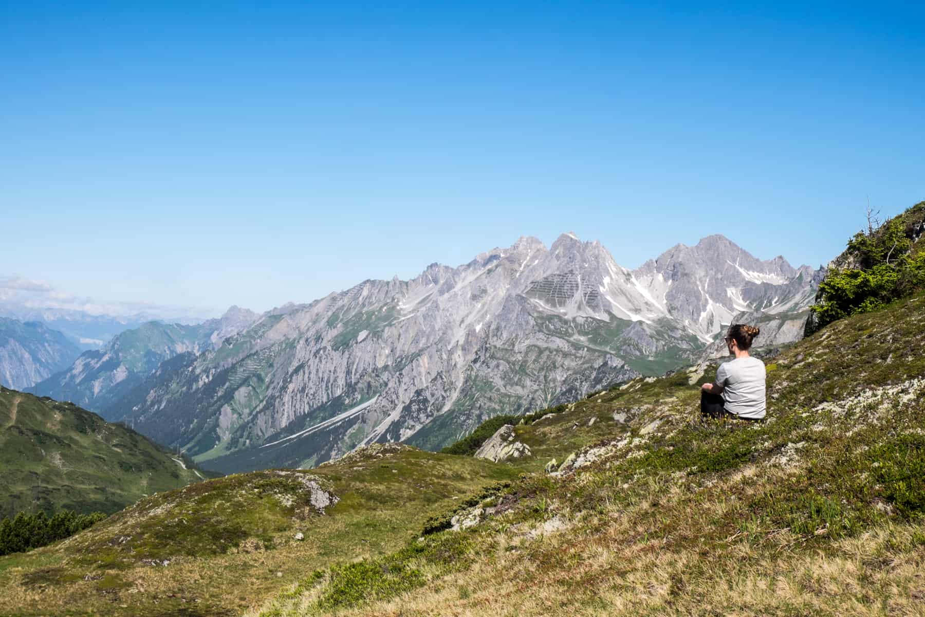 A woman sits on rocky green area in St Anton in Austria, that overlooks a silver mountain peak in the distance