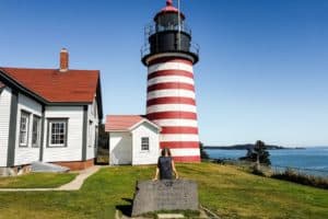 A woman stands in front of a red and white stripped lighthouse on manicured green grass next to the ocean.