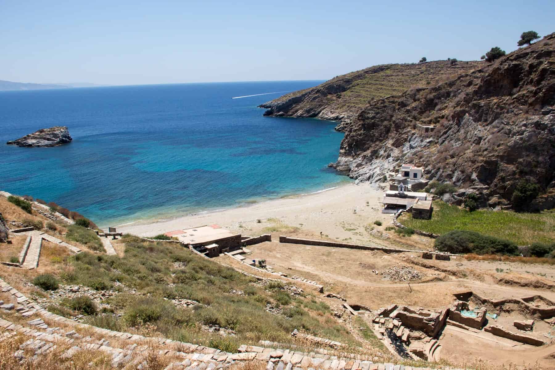 A rugged path and scattered fragments of ruins of the ancient city of Kea leads down a hill to a white sandy beach cove below