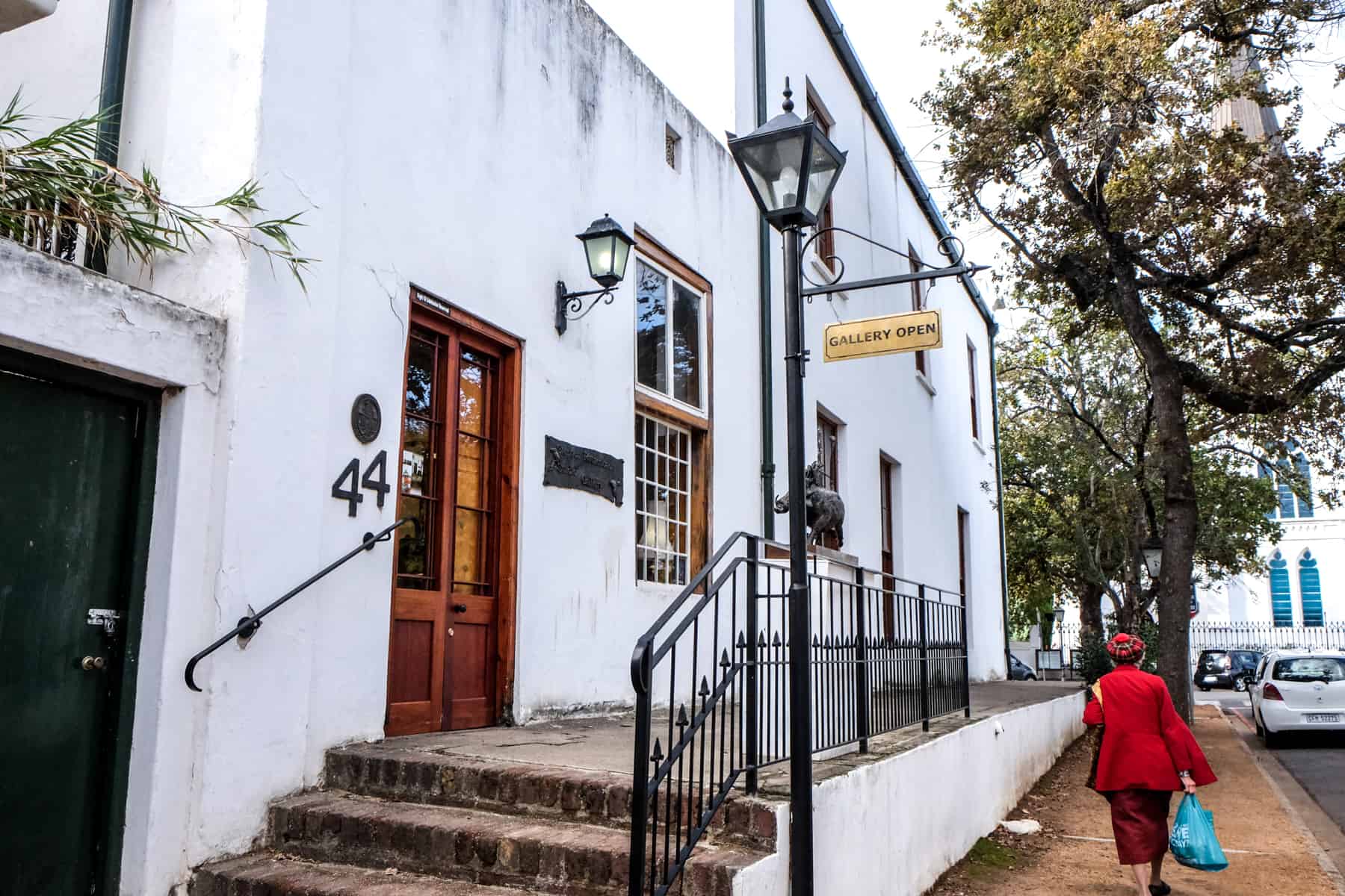 A woman wearing red walks past a white buildings with a red door and brown steps in Stellenbosch