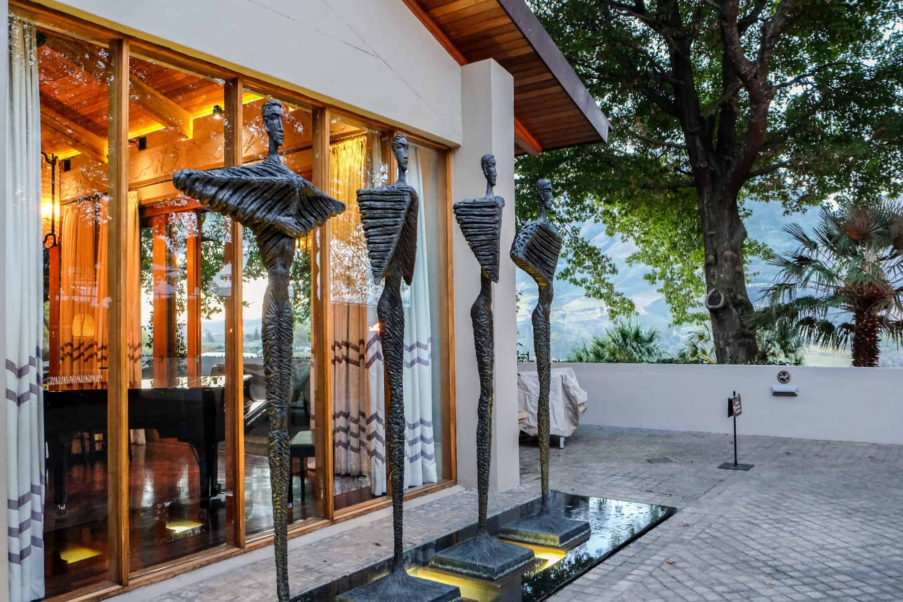 Four metal statues that resemble angels with wings stand outside a white building with a golden lit interior and next to grand trees. They are artworks on display at the Delaire Graff Estate in Stellenbosch 