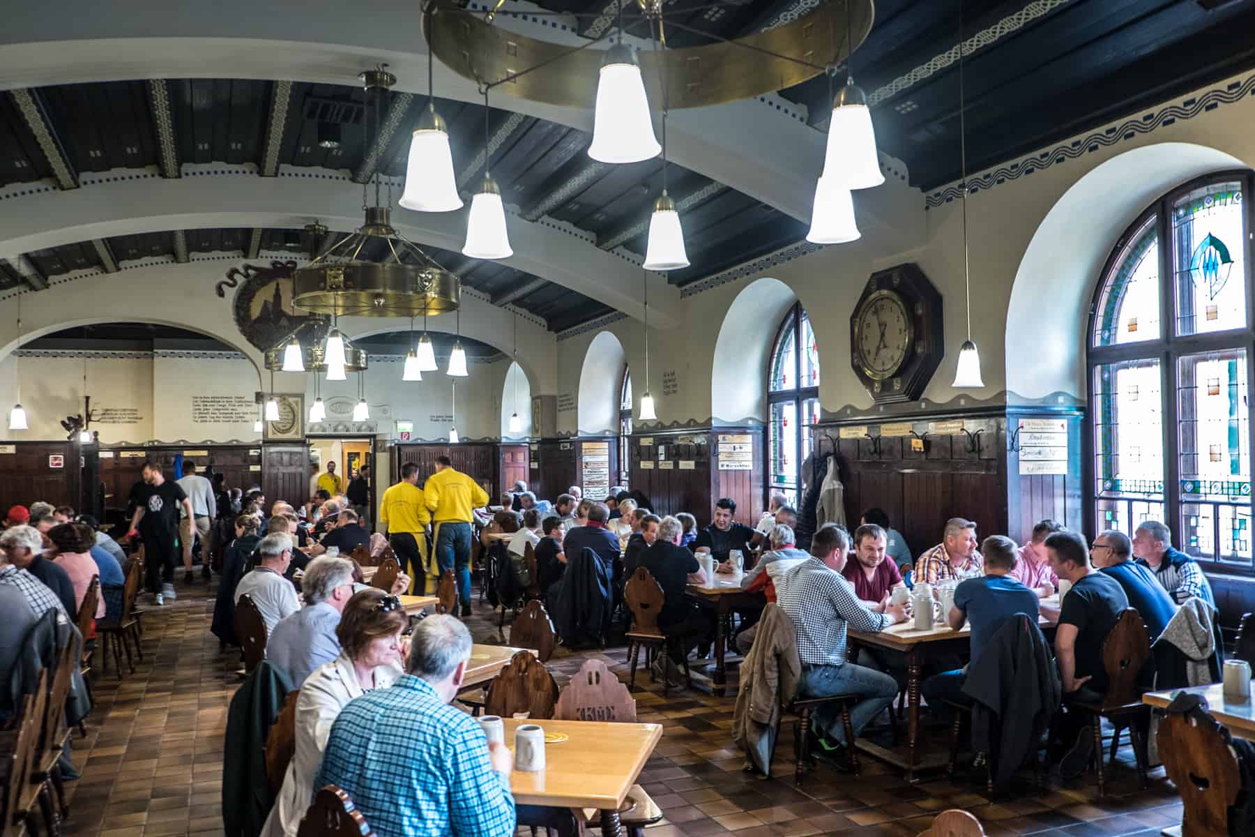 Dozens of people gather around large tables, holding tall ceramic mugs of beer in a huge hall with classic white and dark wood interior.