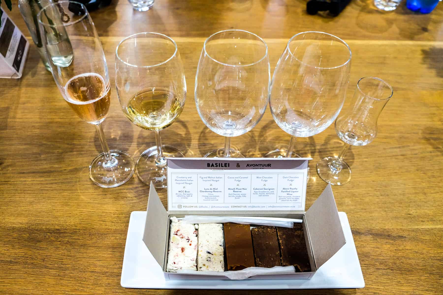 A white box of 5 assorted fudge flavours in front of a row of glasses - one champagne class, three wine glasses and one liquor glass - part of the Avontuur Estate Wine and Fudge Pairing in Stellenbosch 
