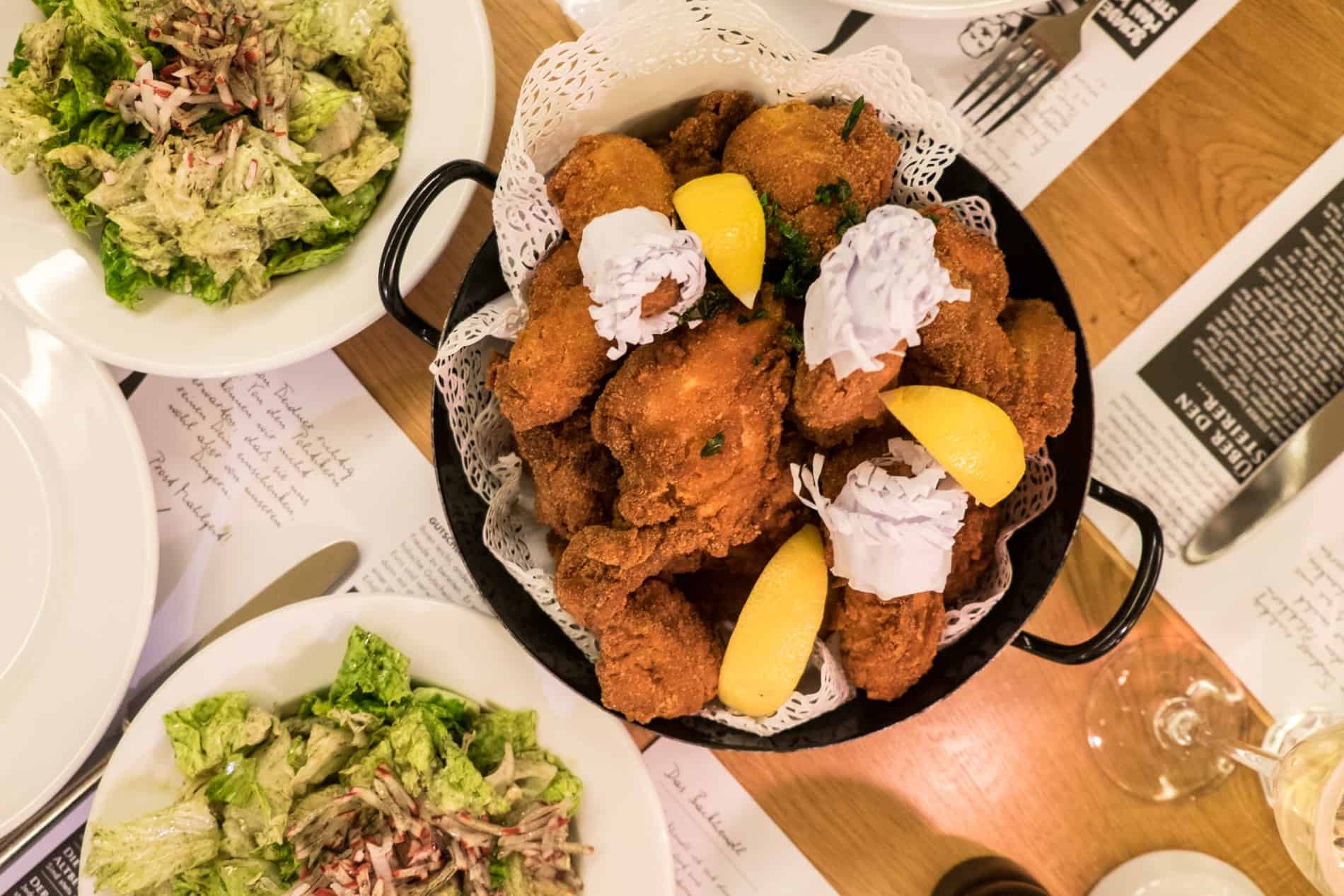 A dish of breadcrumbed chicken with lemon slices and two plates of green salad - Backhendl salad served at The Steirer in Graz.