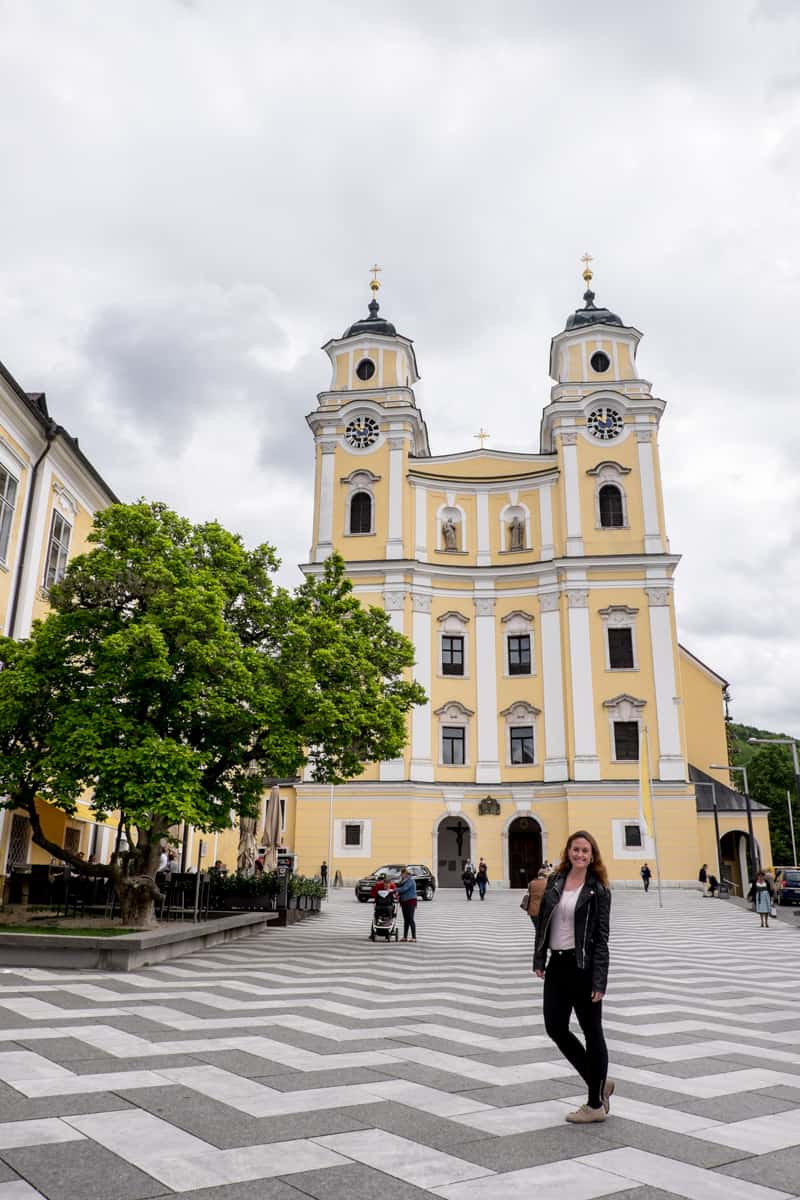 A woman dressed in black stands on the jagged white and grey pavement outside of the yellow and white, two-towered Mondsee Cathedral in Austria. 
