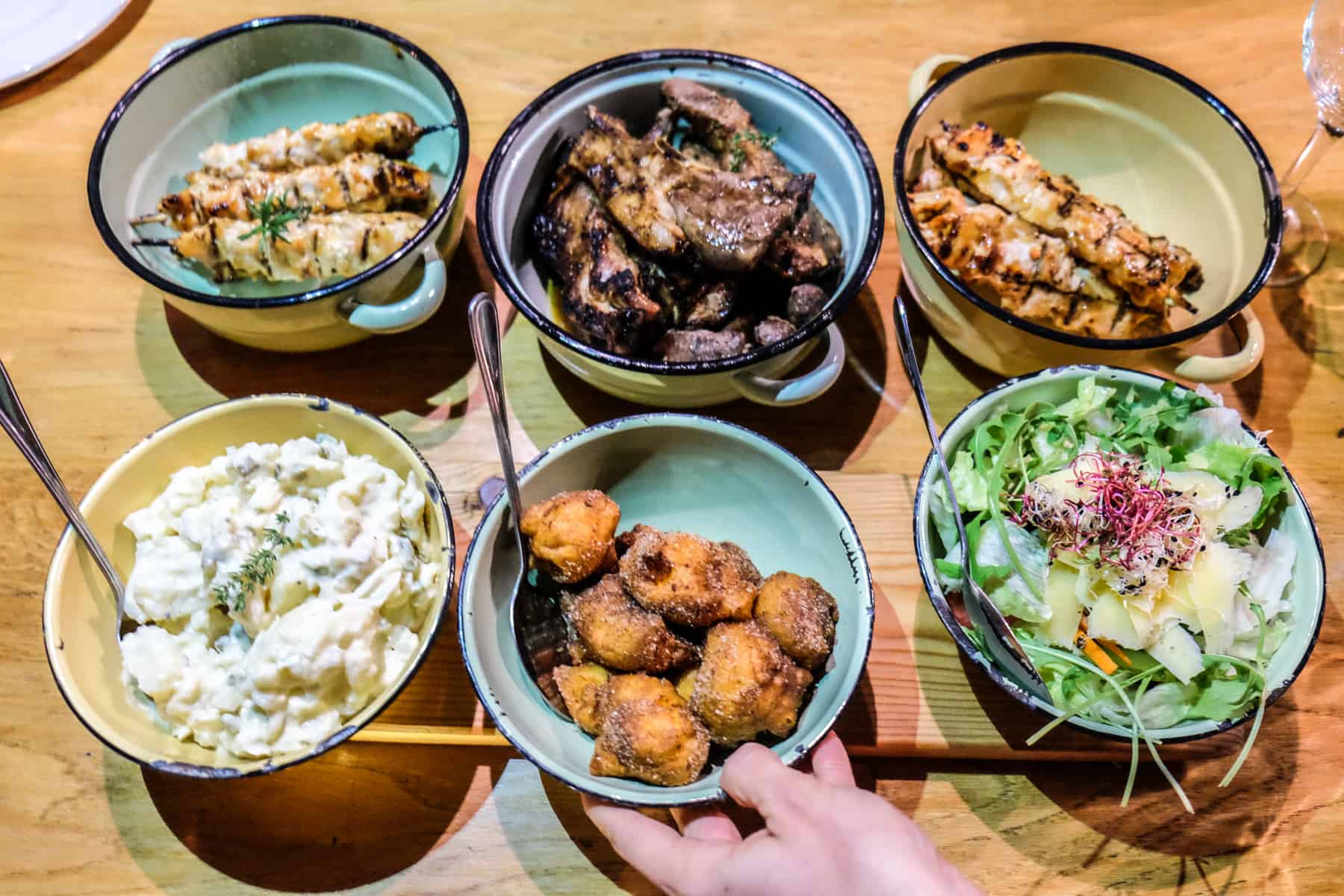 6 bowls of meats, sauces and fried goods that make up a typical South African Braai. A hand is shown lifting the middle bowl on the bottom row