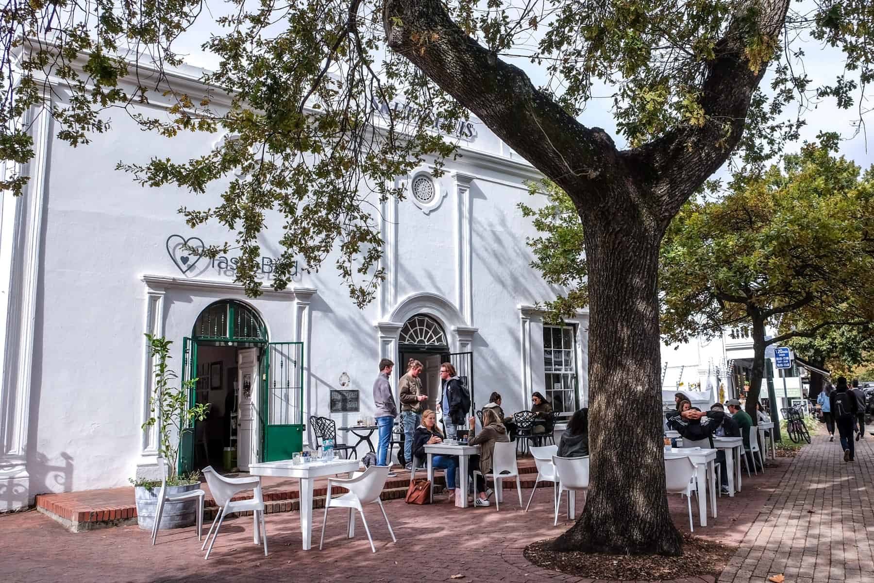 A group of people gather at a cafe in Stellenbosch, set within an old classic white building behind a giant tree