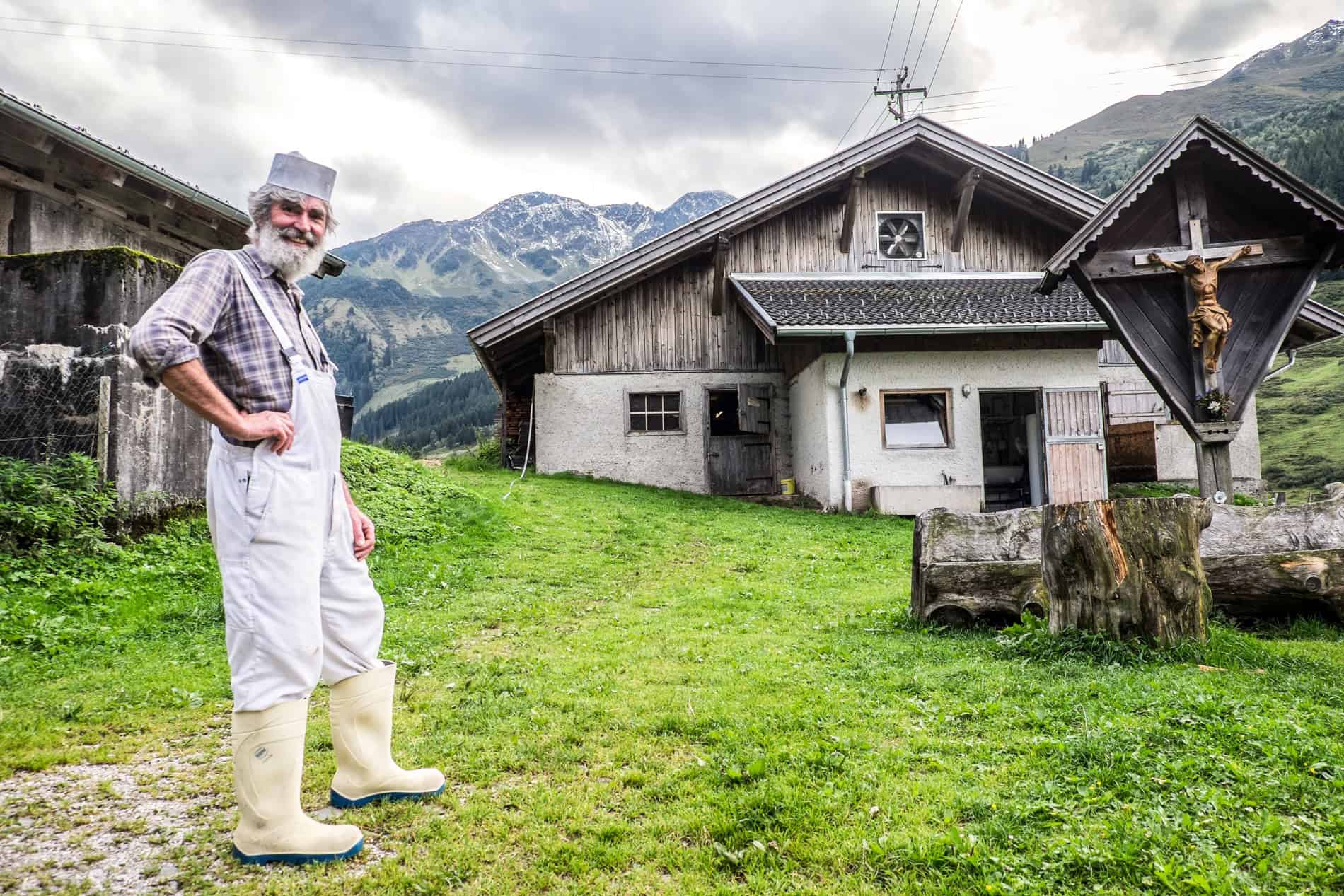 A man in white overalls , with a white hat and beard, stands on a green lawn outside an alpine hut in Austria. 