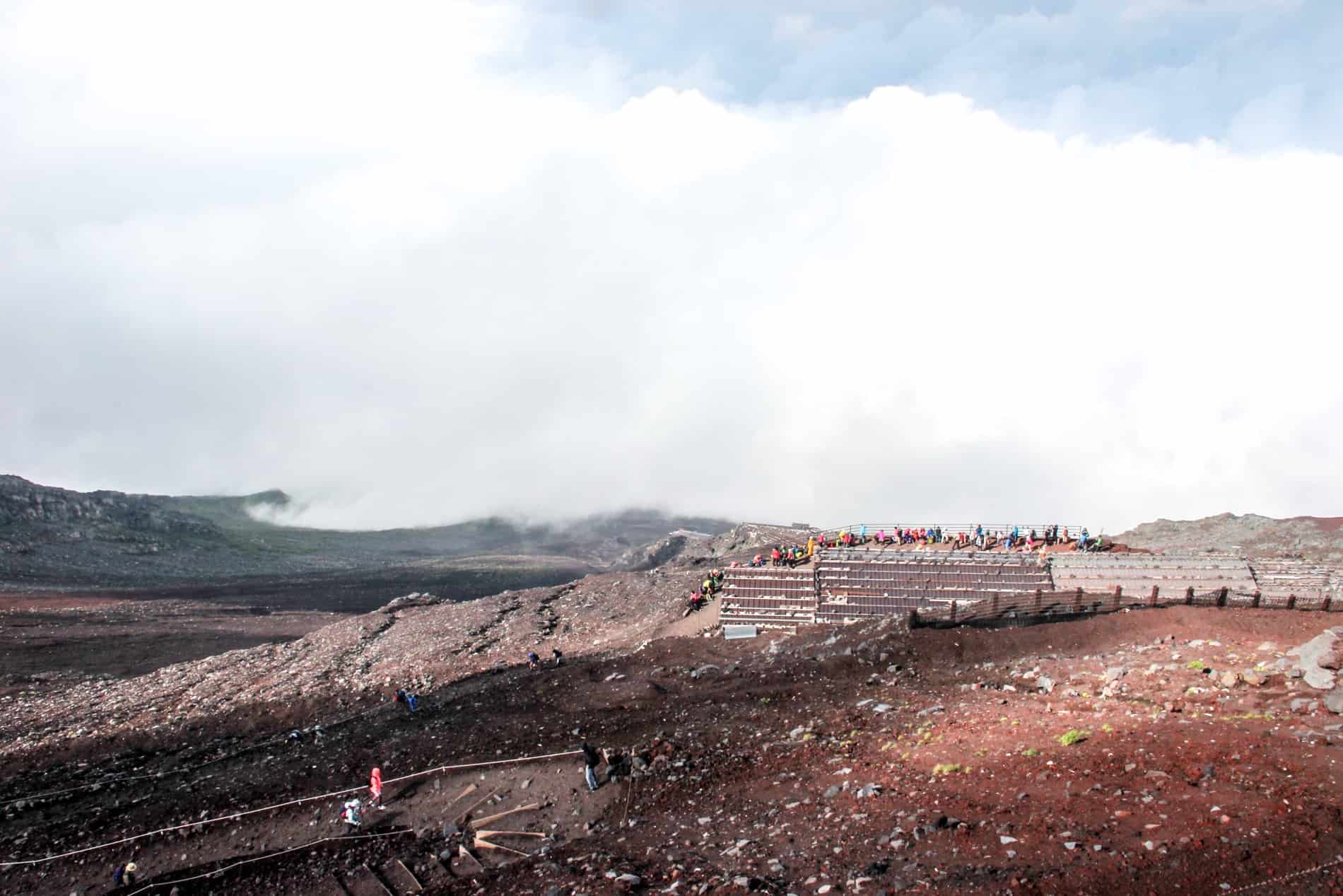 Hikers wearing colourful gear at an elevated rest point on the red volcanic rock of Mt. Fuji. 