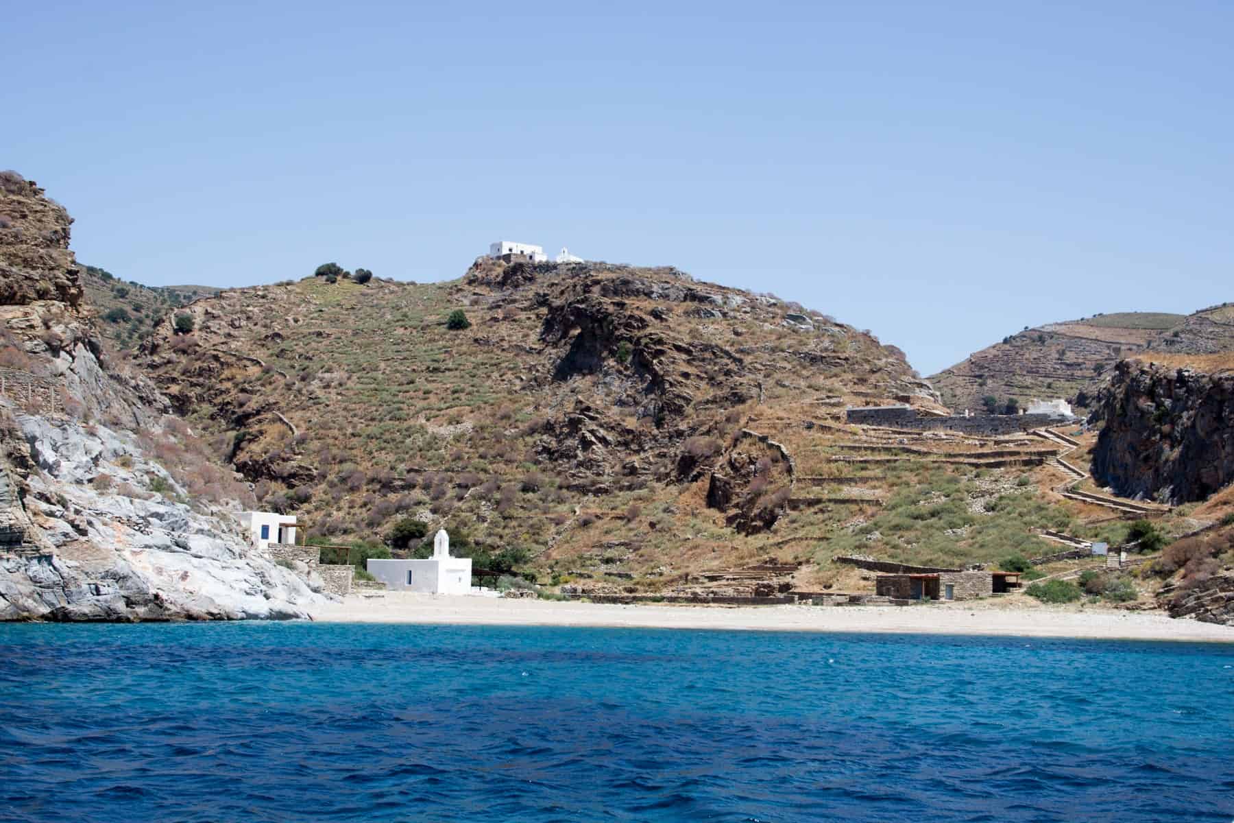 A far view of a sandy cove on Kea island marked by white buildings and the lines of a walking trail up to ancient monuments on the hillside