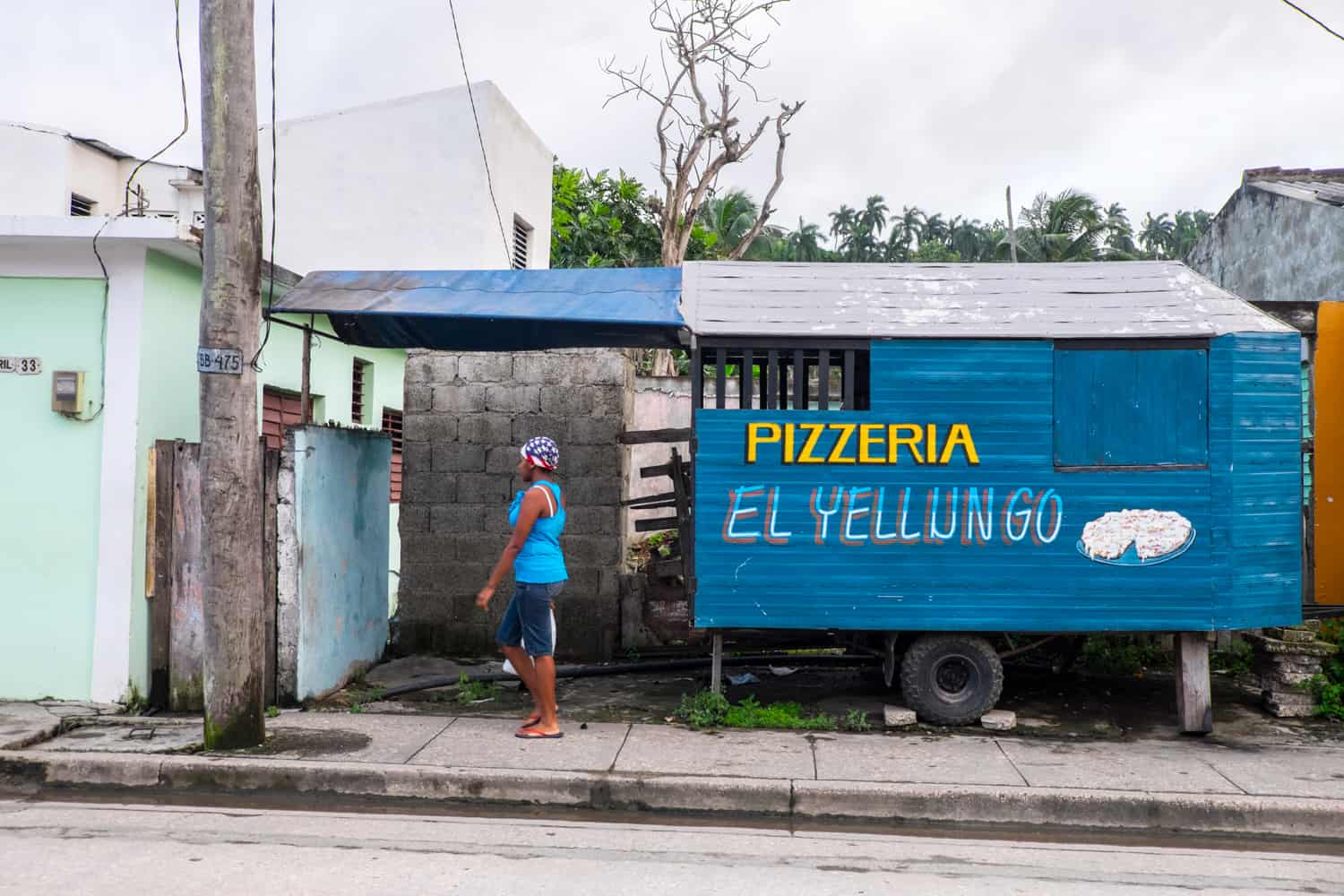 In Cuba, a woman with an American flag head bandana walks past a blue wooden shack with the words: Pizzeria El Yellungo